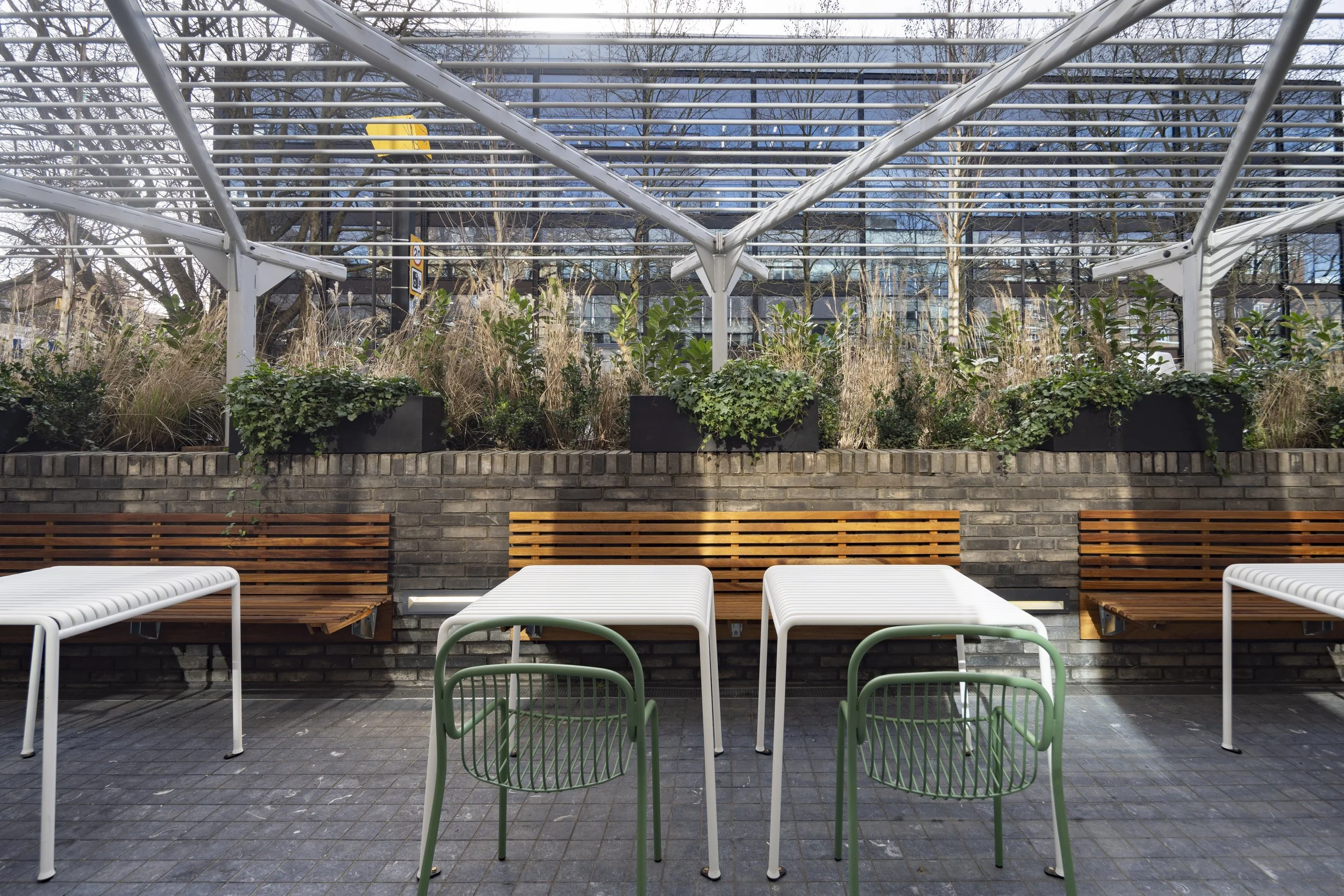 Empty outdoor patio with white tables, green and wooden chairs, brick wall, and planters with greenery. Metal pergola overhead, city buildings and leafless trees in background.