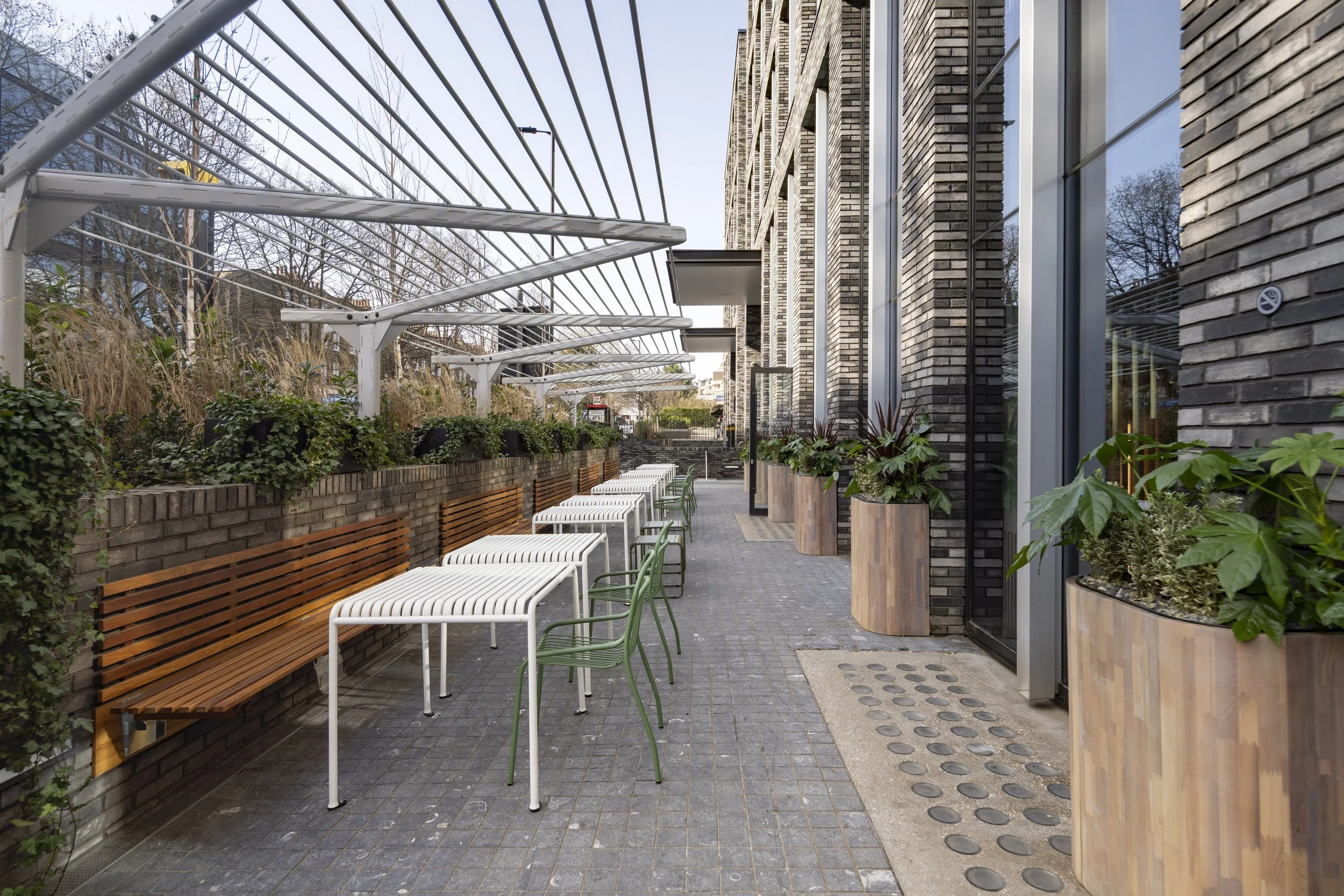 Outdoor patio with white and green chairs, wooden benches, large planters with greenery, brick building, and a clear sky.