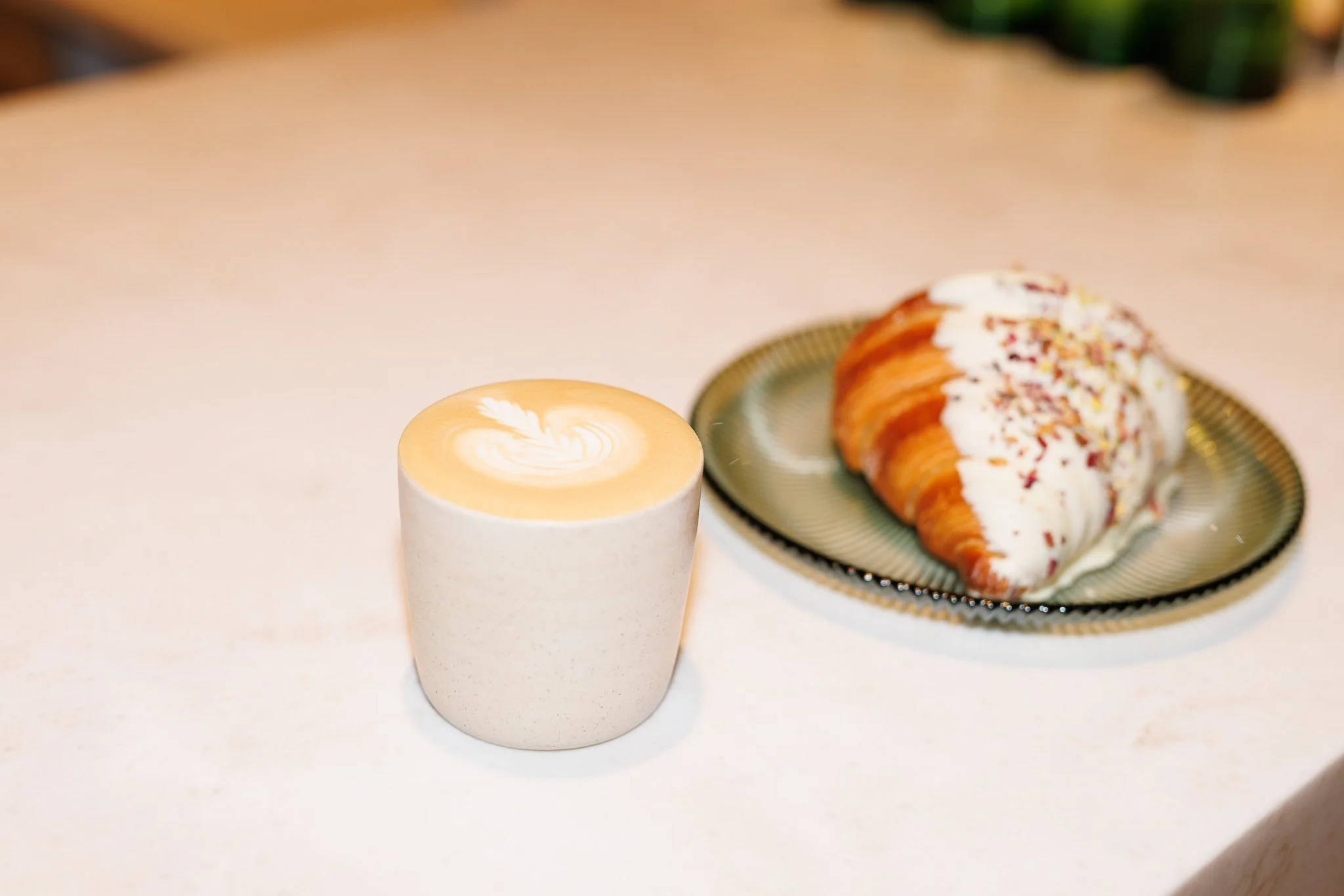 A beige coffee cup with a latte art design on top, and a plate with a pastry topped with icing and sprinkles, on a white surface.