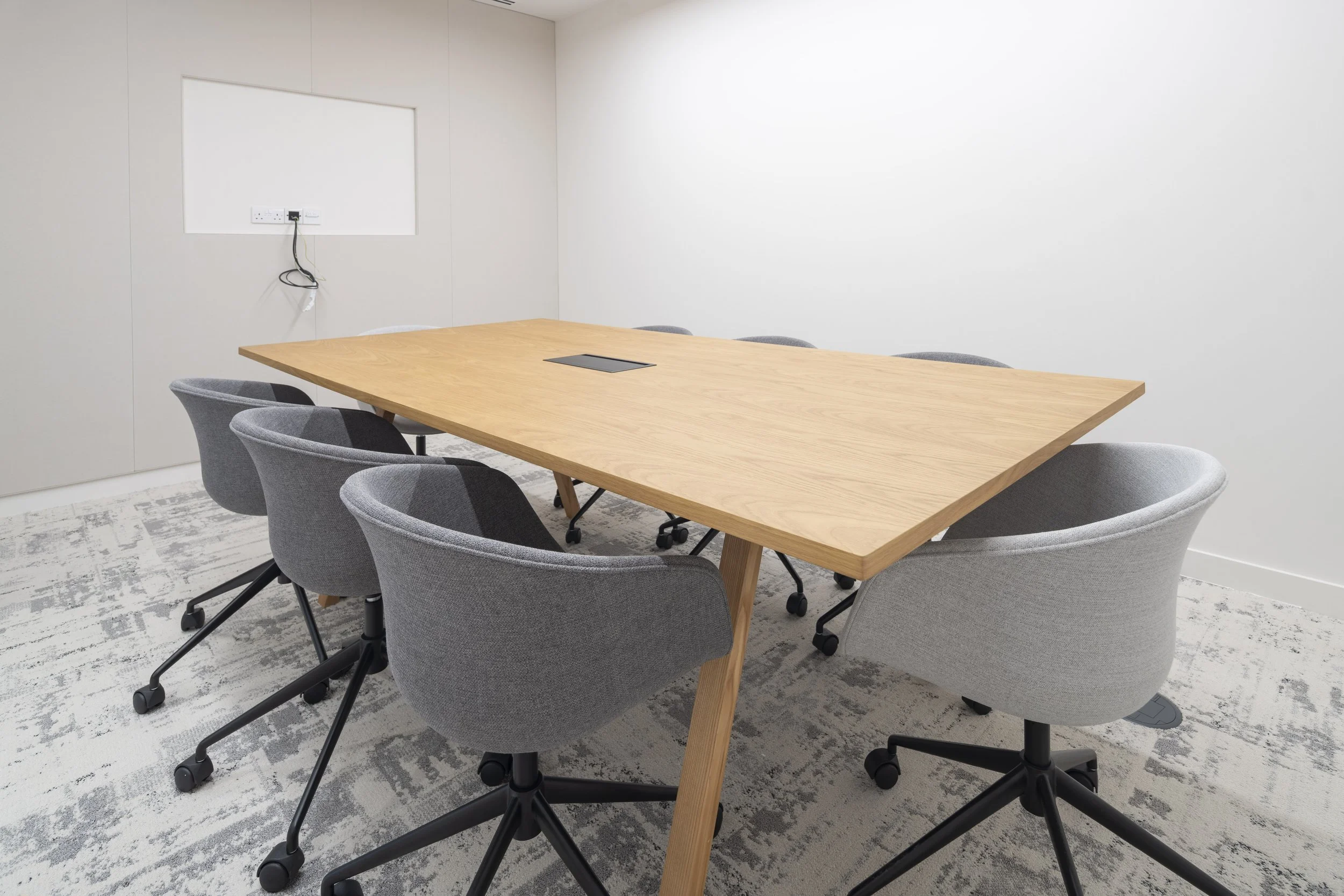 Empty conference room with a wooden table and eight gray chairs, set against plain white walls and a partially visible patterned carpet.