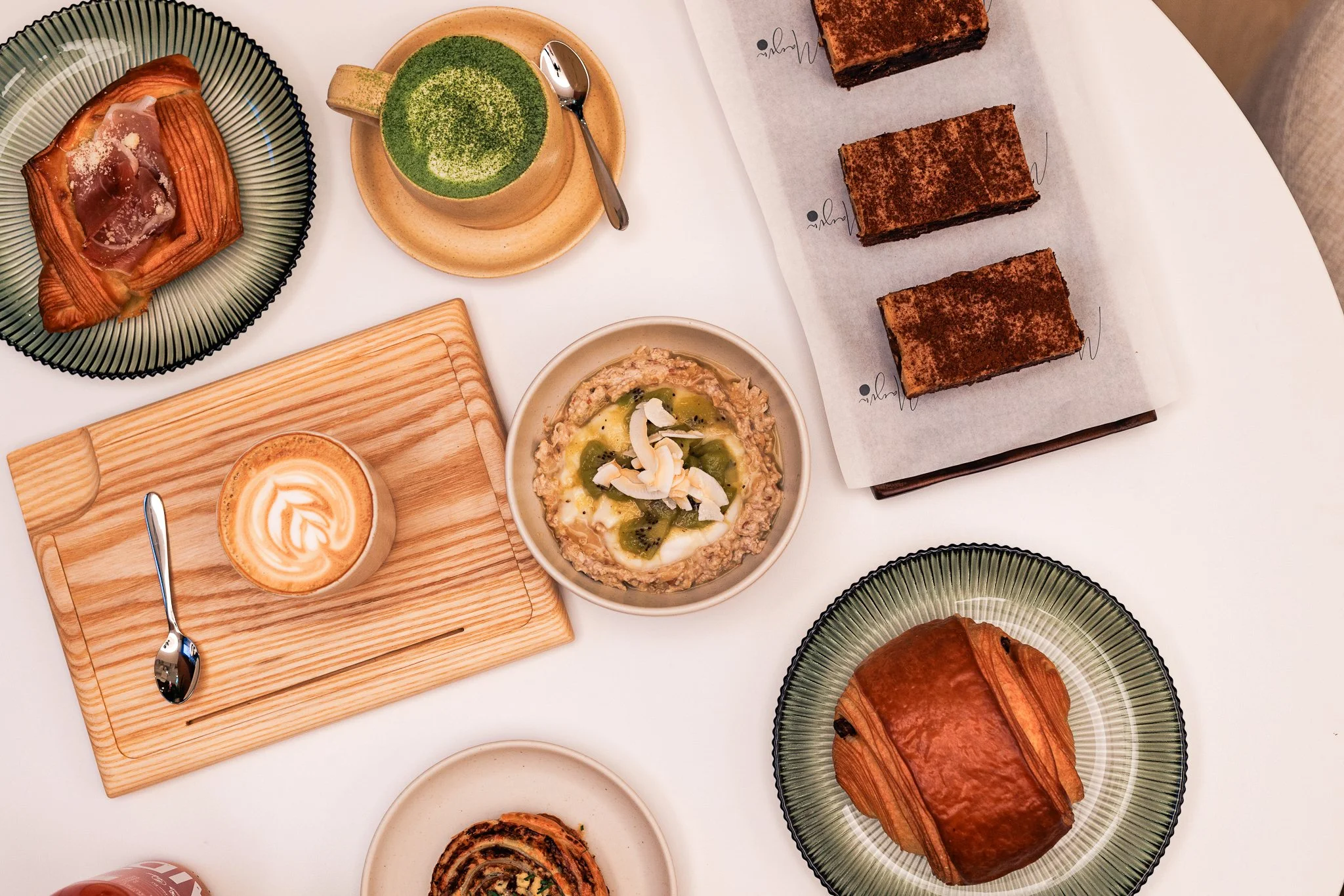 A table with various desserts and drinks including a croissant with ham, a matcha latte, a cappuccino, a bowl of oatmeal topped with coconut flakes and kiwi slices, and three pieces of tiramisu. There are also two plates with croissants and a spiral 