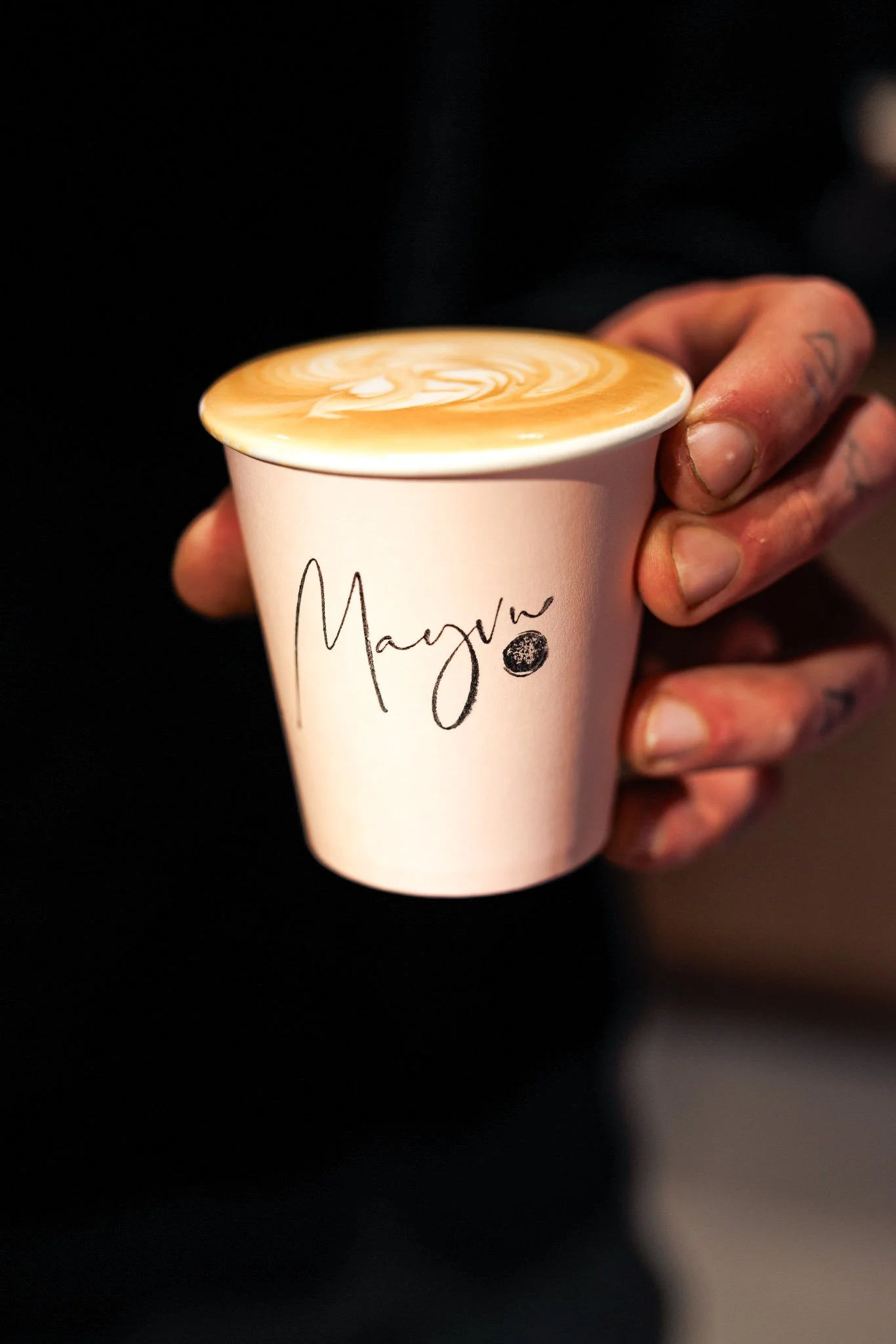 A person with tattooed fingers holding a paper coffee cup with a latte that has latte art on top. The cup has the word 'MAYEVE' written on it.