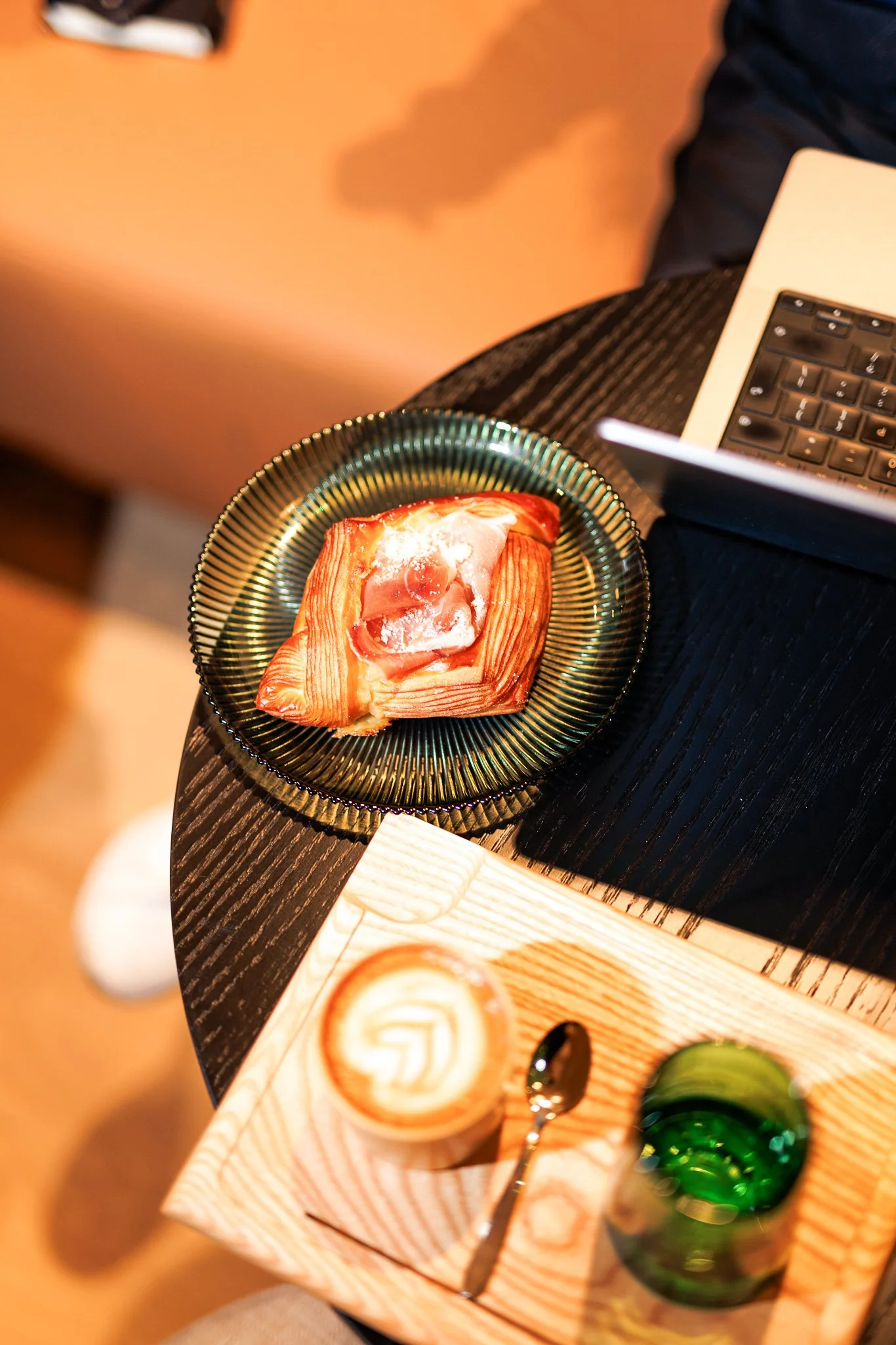 A piece of smoked salmon with sliced onions on top, placed on a decorative black plate. A bowl of ramen, a green glass, a spoon, and a cup of coffee with latte art are on a wooden serving board nearby.