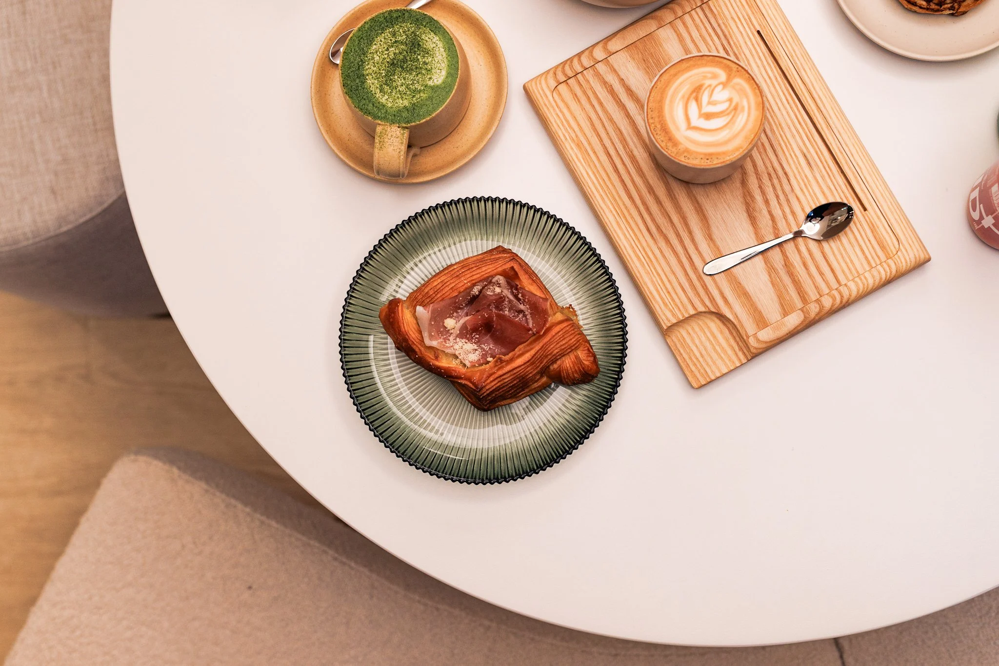 A round white table with a green matcha latte, an orange latte with foam art, a pastry with ham on a green and black striped plate, and a cup of coffee on a wooden tray.