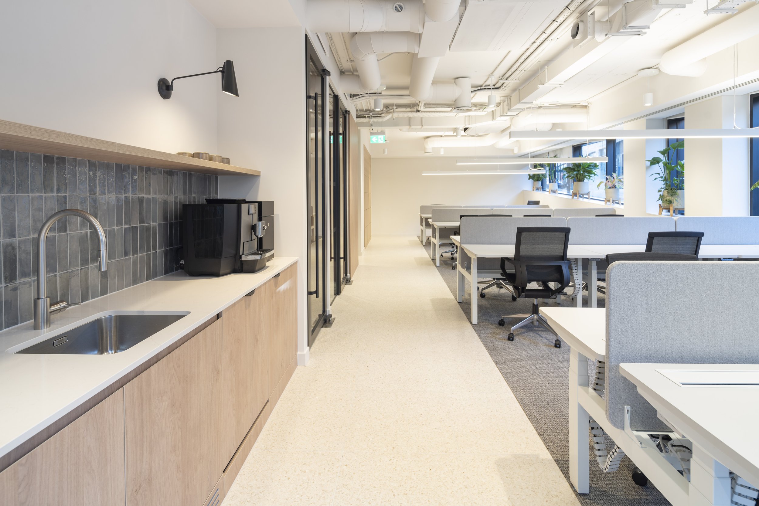 Modern office break area with a coffee machine, sink, and countertop on the left, and workstations with chairs and gray privacy screens on the right, illuminated by natural light from large windows with potted plants.