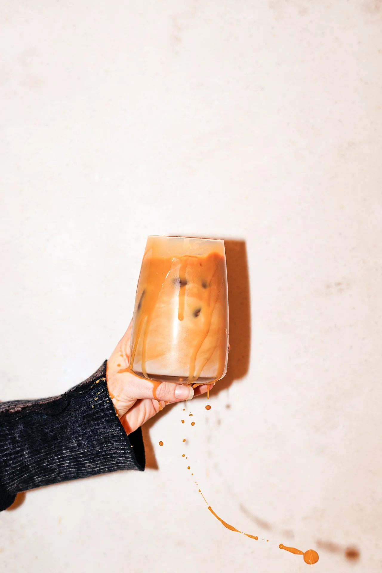 Hand holding a glass of iced coffee with caramel drizzle, spilling some of the drink against a plain light-colored wall.