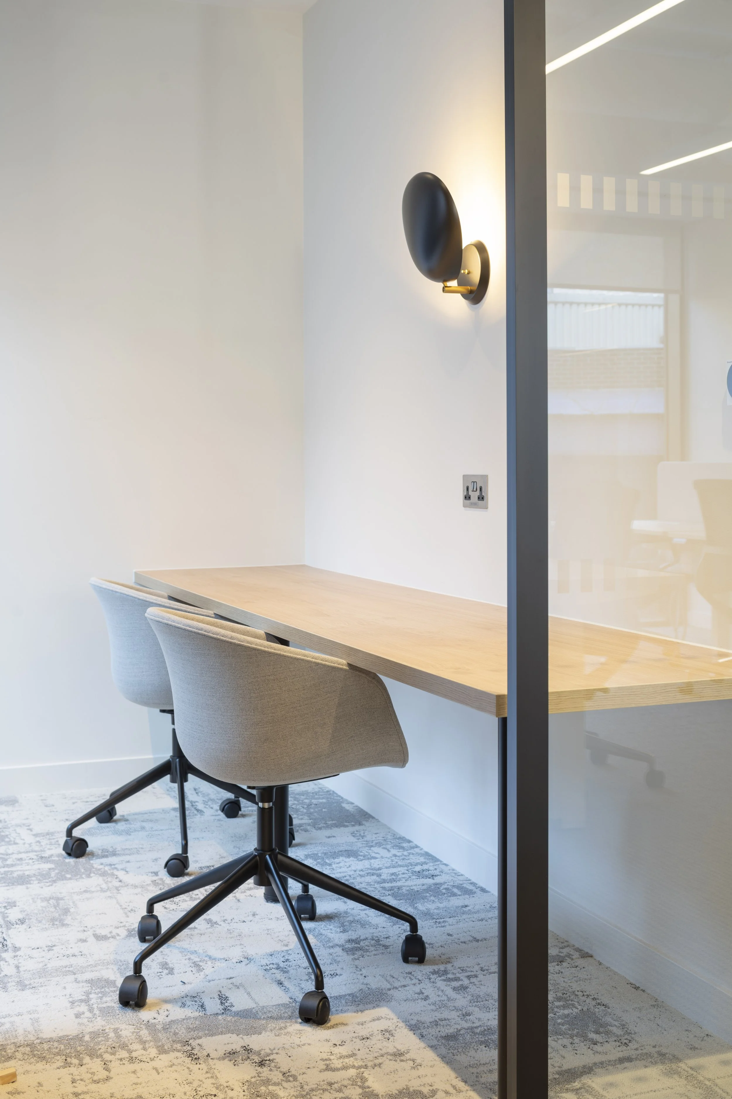 An empty office workspace with a wooden table, two beige chairs, a wall-mounted black and gold light, and a power outlet on the wall. Part of a glass partition is visible on the right.