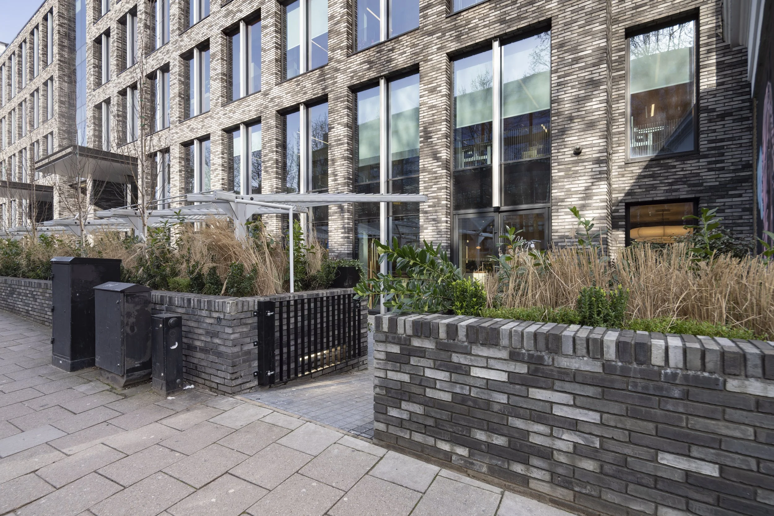 Modern urban building with large windows, brick exterior, small garden area with plants, bushes, and a brick fence, sidewalk in foreground.