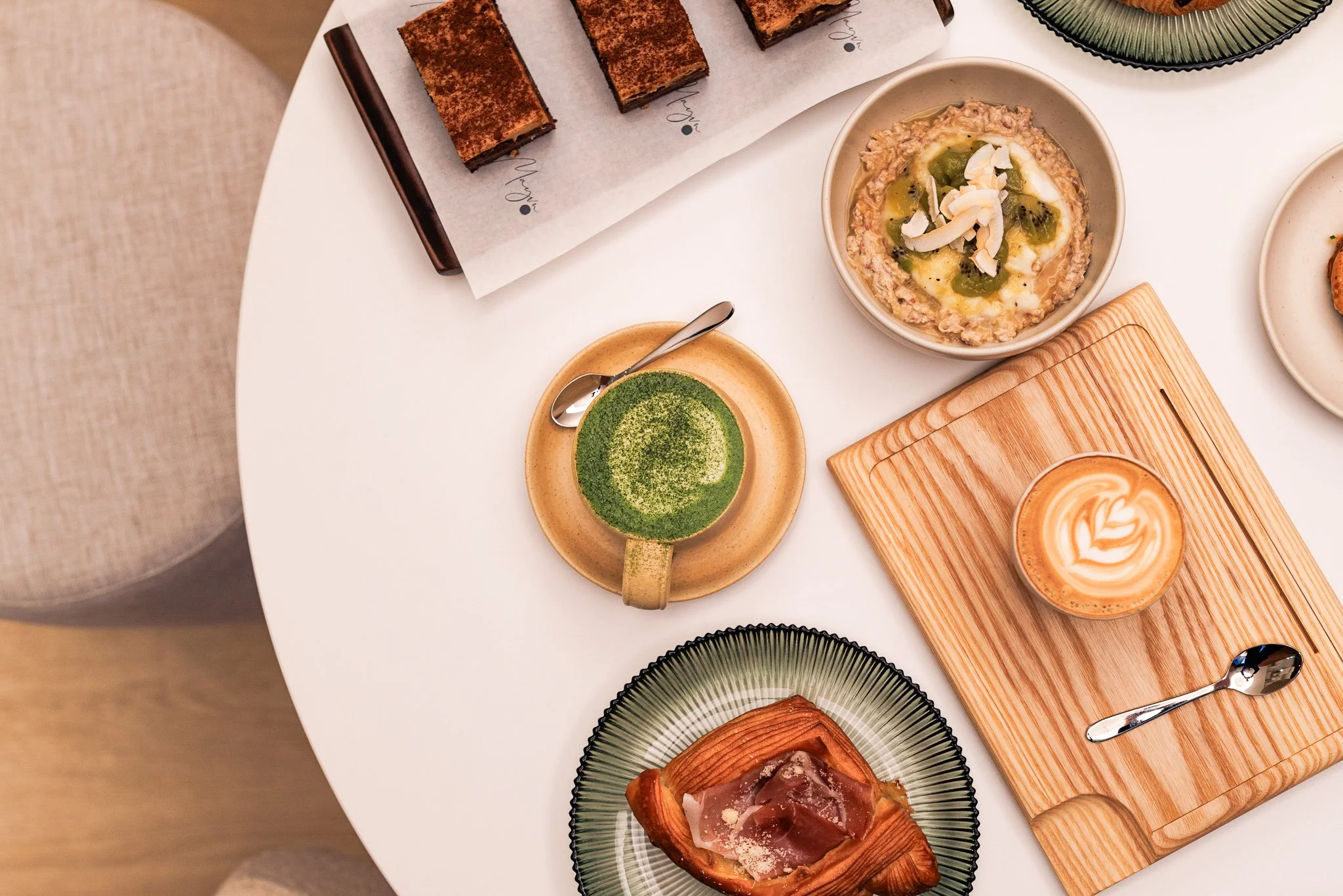 A top-down view of a white round table with various desserts and beverages including a plate with a pastry, a green matcha latte in a ceramic cup, a latte with foam art in a glass on a wooden board, a bowl of porridge topped with coconut flakes and f
