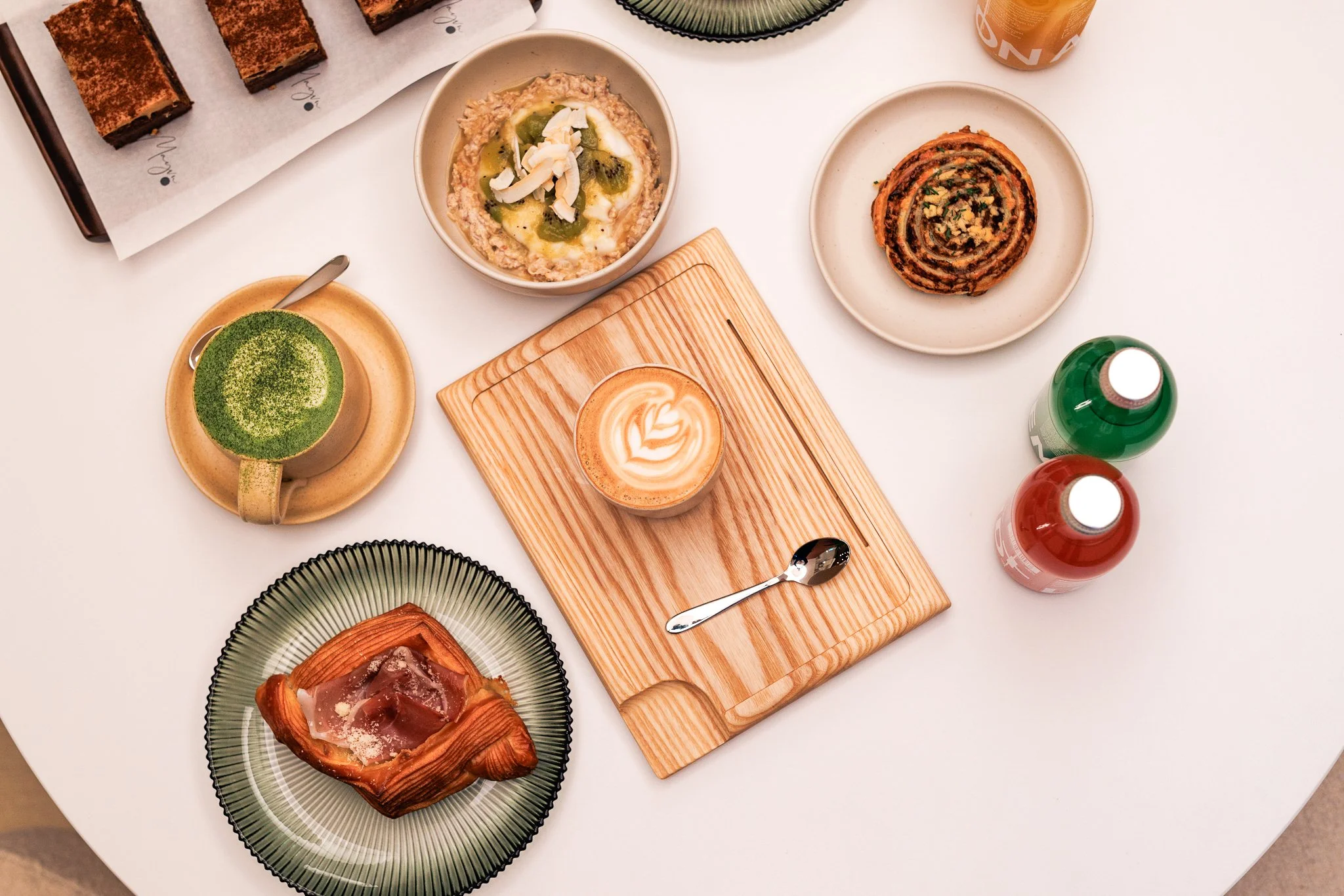 A top-down view of various desserts and beverages on a white table, including a matcha latte, a coffee with latte art, a cinnamon roll, a pastry, and bottled drinks.