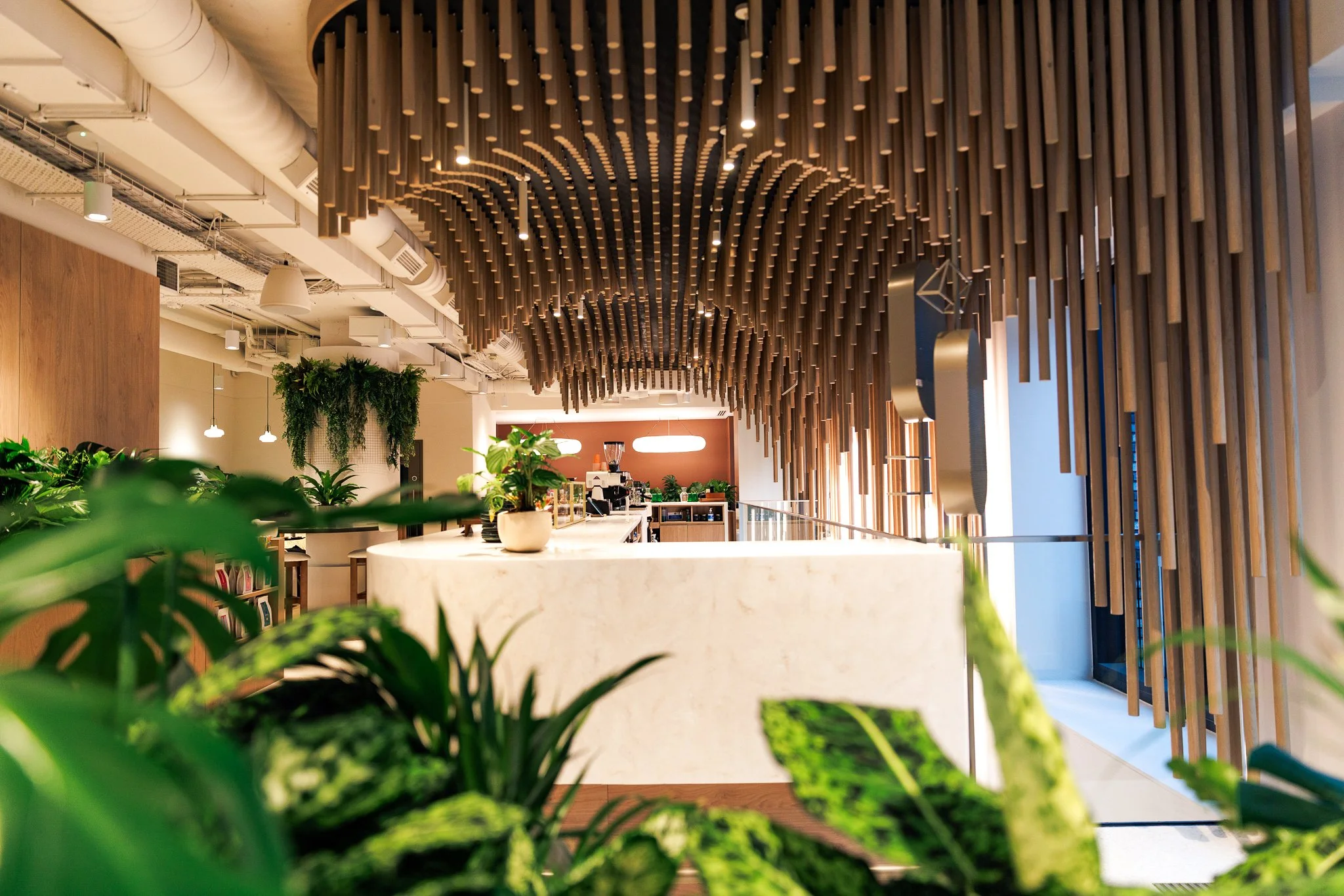 Interior of a modern cafe or restaurant with a white marble counter, wooden vertical slats on the ceiling and wall, green plants, and warm lighting.