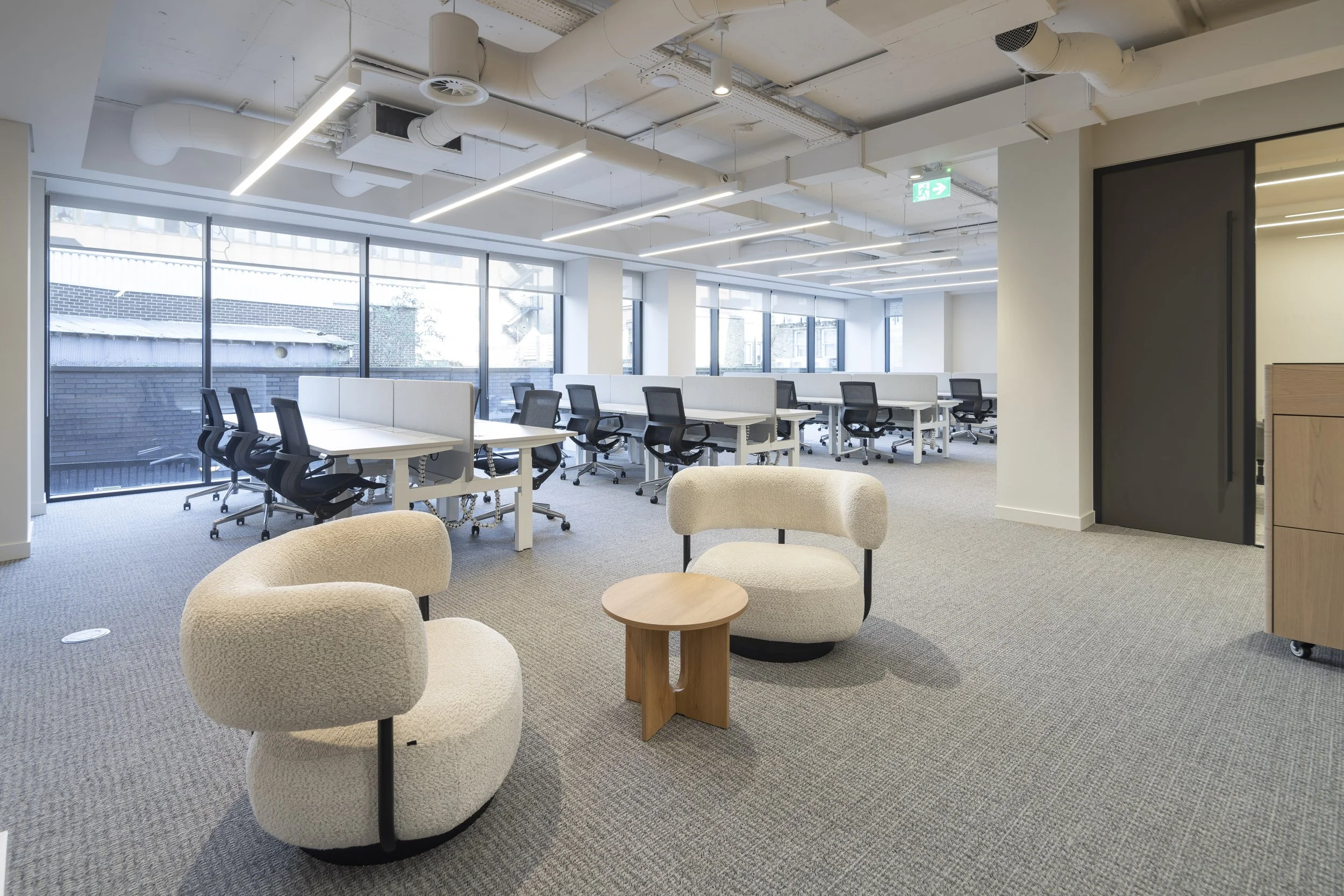 Modern, open-plan office space with large windows, white desks, black office chairs, and two beige, rounded lounge chairs with a small wooden table between them, carpeted flooring, and exposed ceiling ducts and lighting fixtures.