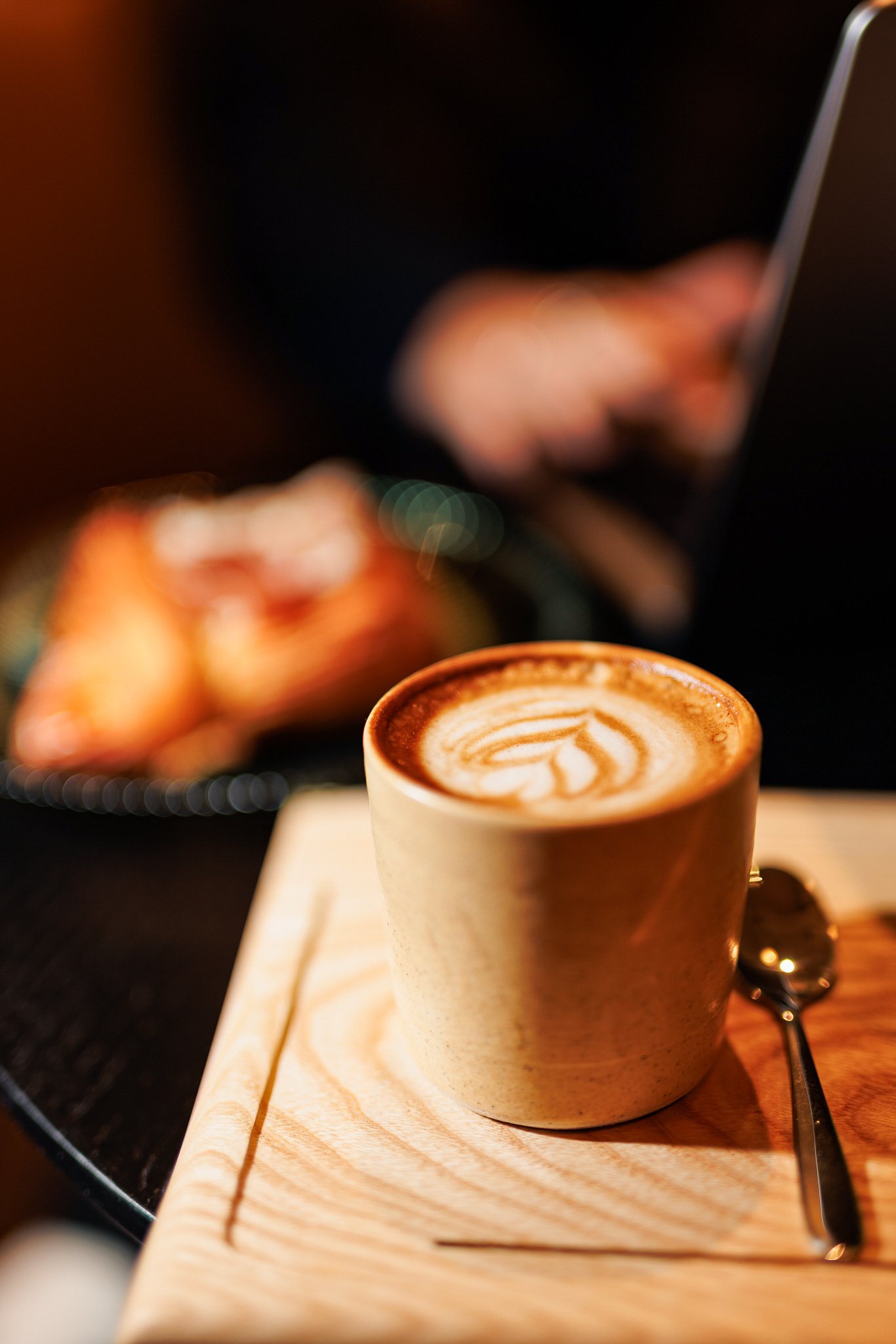 A latte with foam art in a beige mug on a wooden tray with a spoon, with a blurred plate of croissants in the background.