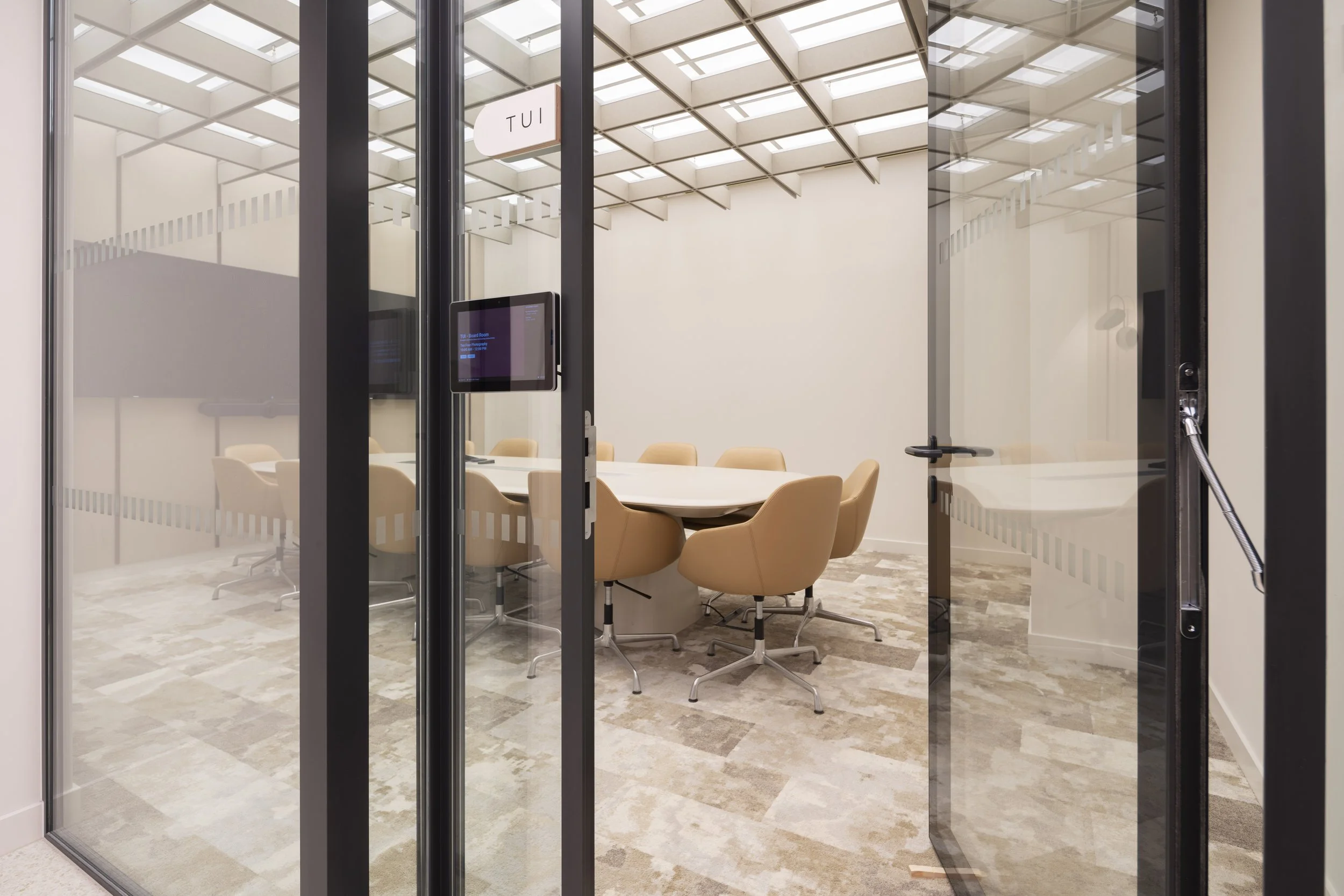 Empty modern conference room with glass walls, beige chairs around a circular table, and a wall-mounted screen.