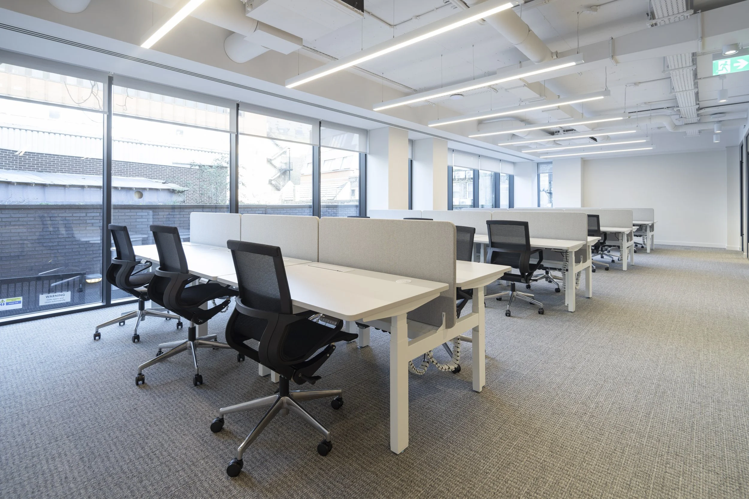 Empty modern office space with white desks, black office chairs, and large windows with blinds, bright overhead lighting, and gray carpet flooring.