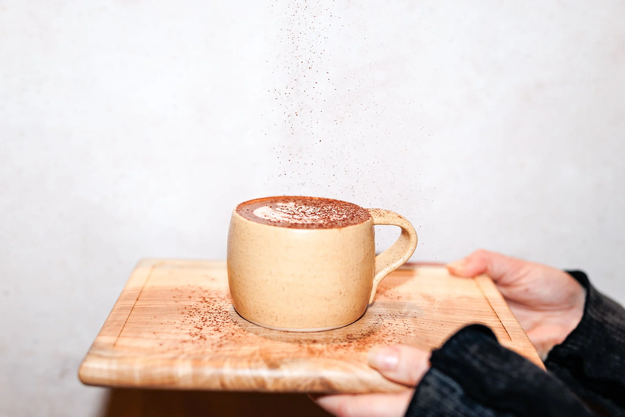 Hand holding a wooden tray with a beige ceramic cup on top, being dusted with cocoa powder