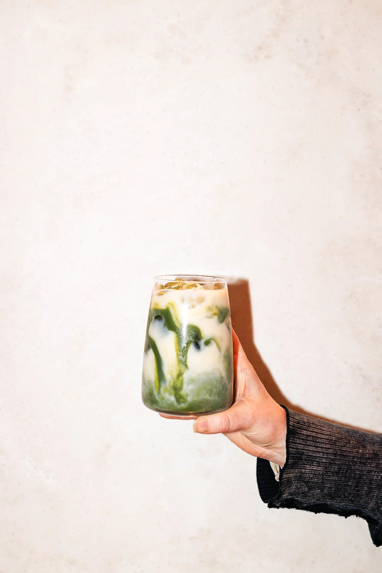A person holding a glass of iced matcha latte with milk, swirled with green matcha and white milk, against a plain light background.