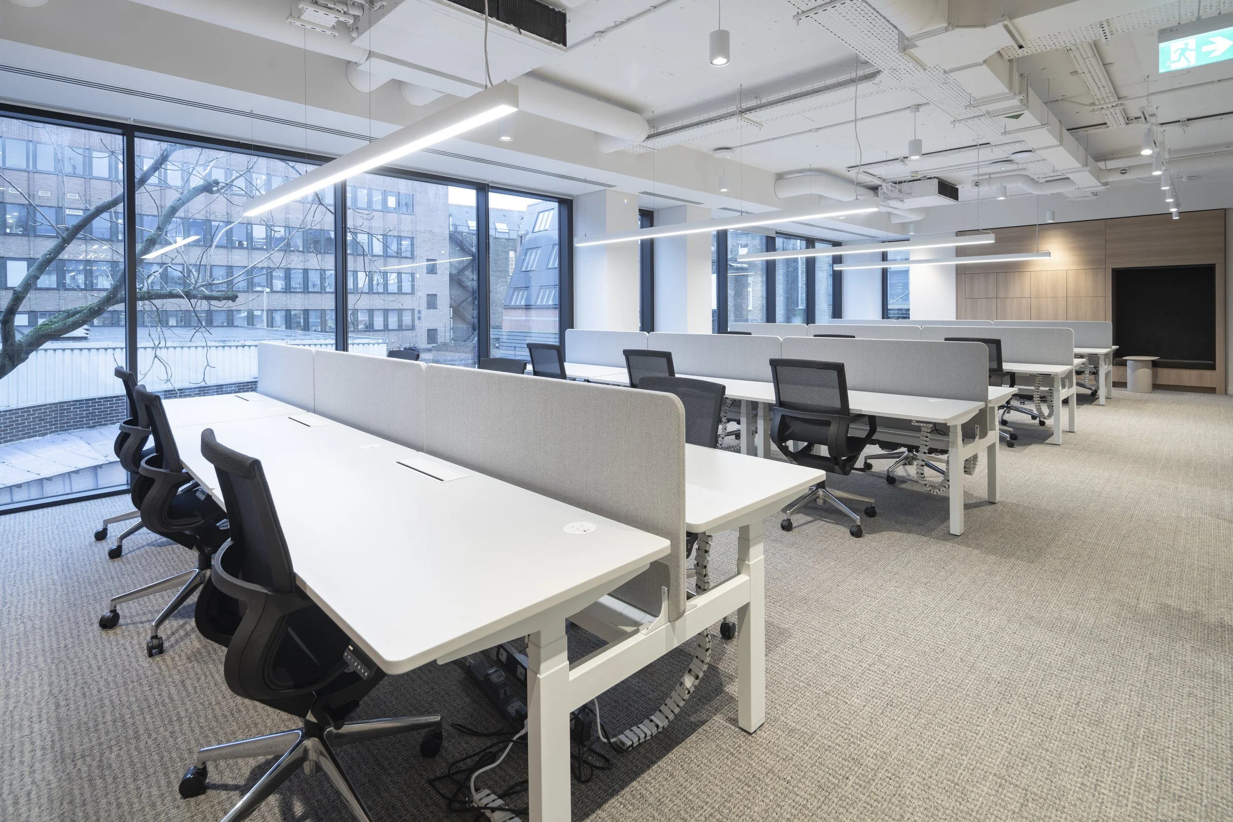 Empty modern office space with white desks, black chairs, and large windows showing an urban exterior, with neutral décor and overhead lighting.