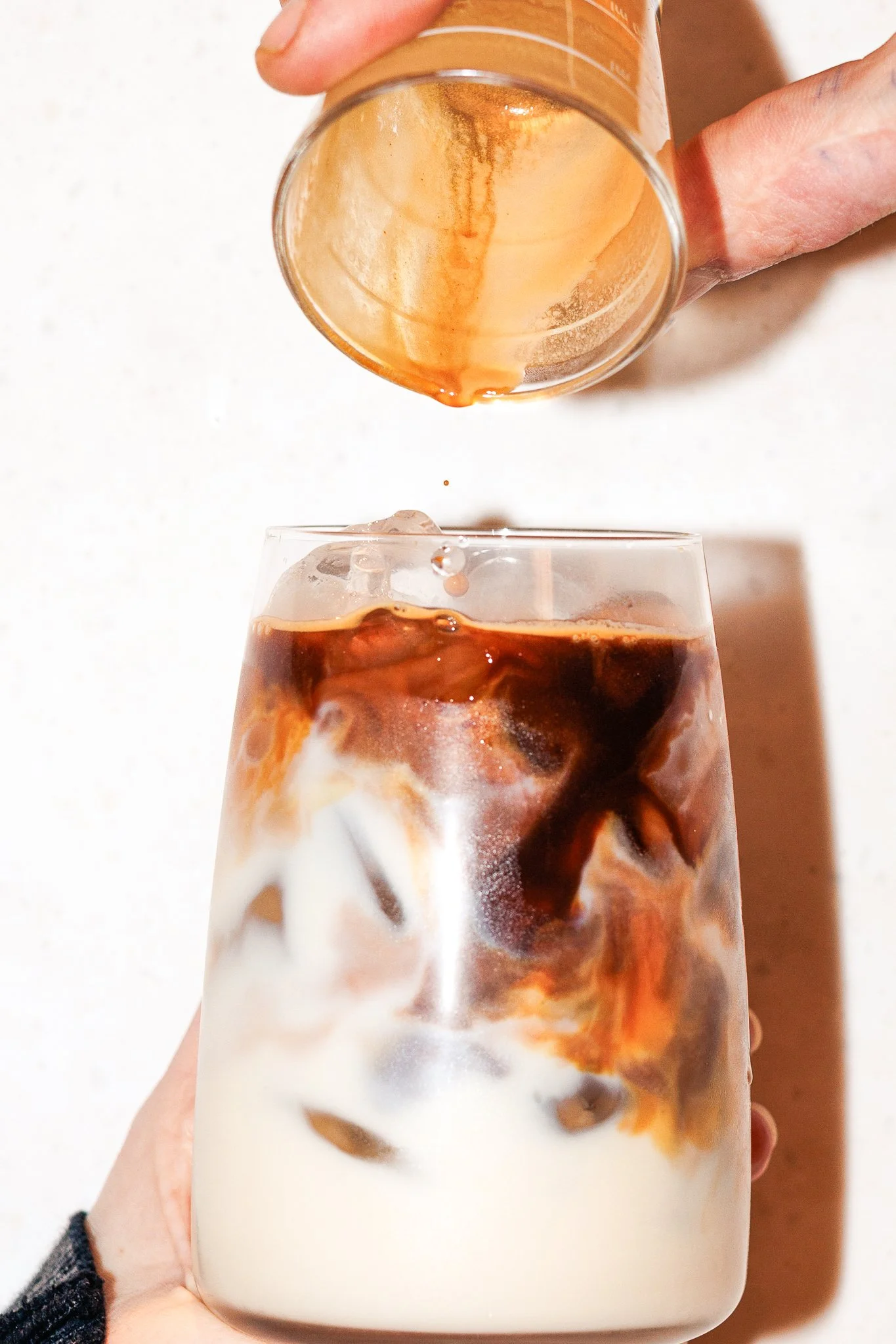 A hand holding a glass of iced coffee or milk tea with ice cubes, with a small container of brown syrup being poured into the glass.