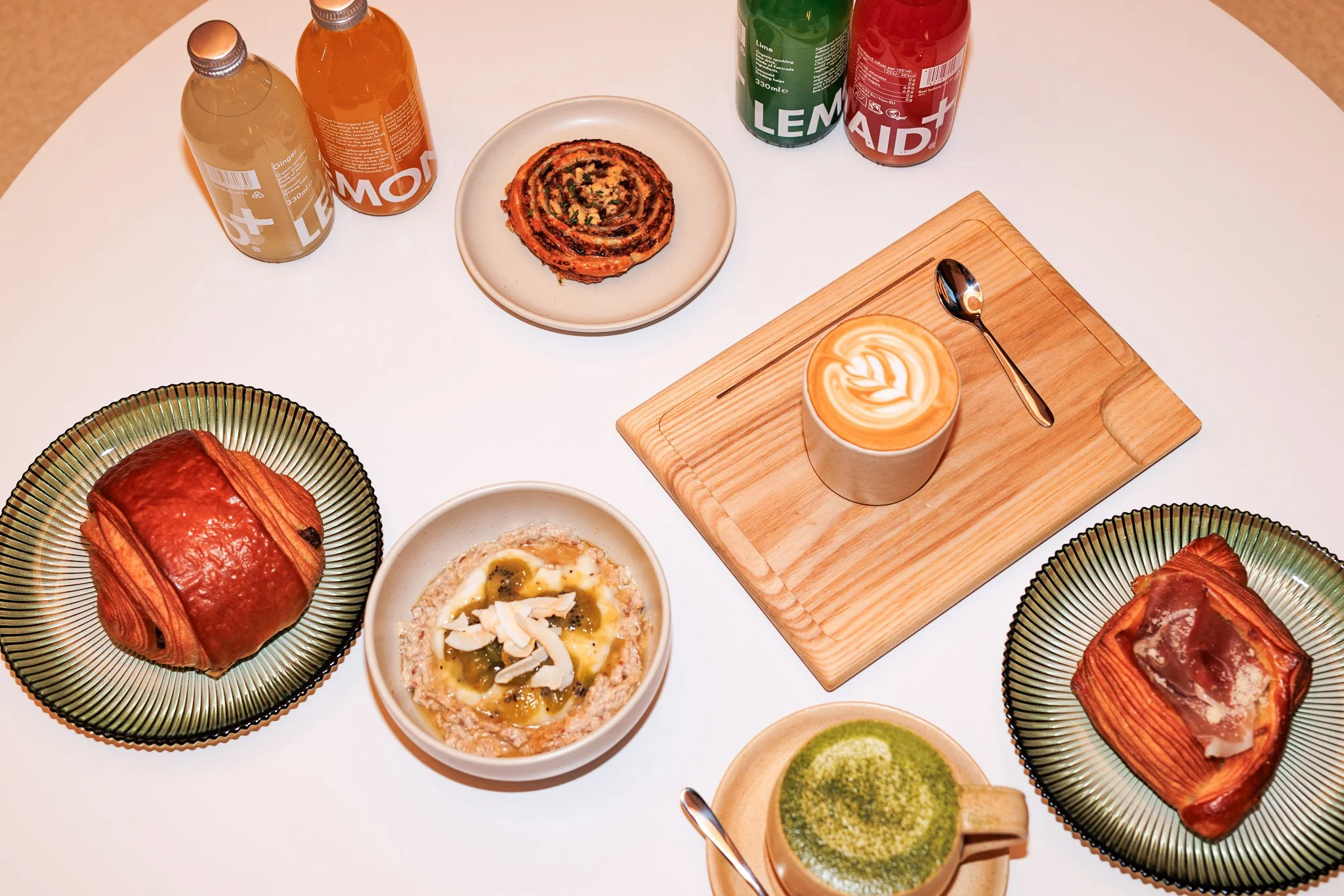 Breakfast table with pastries, beverages, and coffee drinks, including cinnamon roll, pastry topped with ham and cheese, a matcha latte, and a latte with heart-shaped foam art.