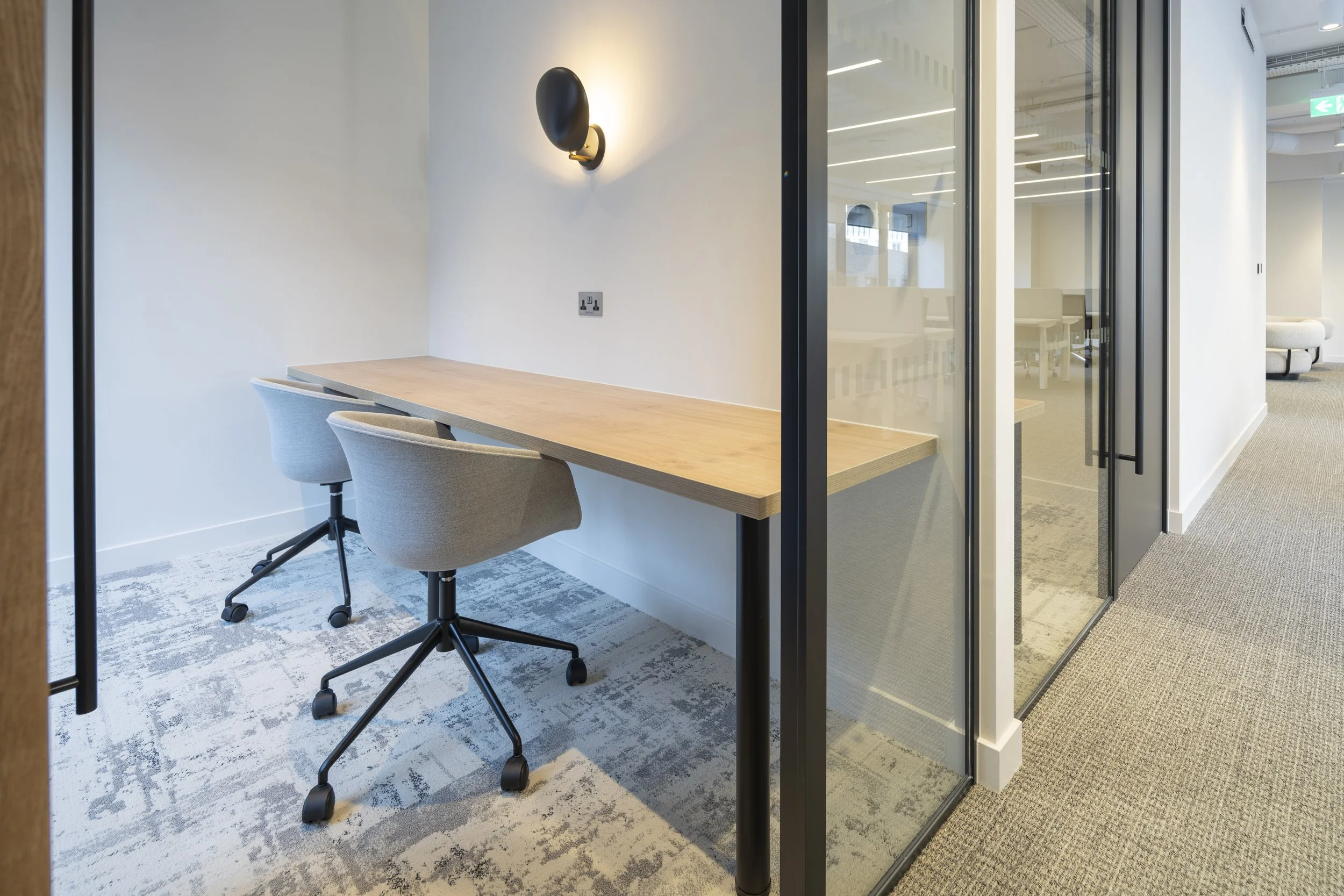 Modern office meeting room with wooden table, two gray swivel chairs, wall-mounted black sconce light, glass wall partition, beige carpet, and white walls.