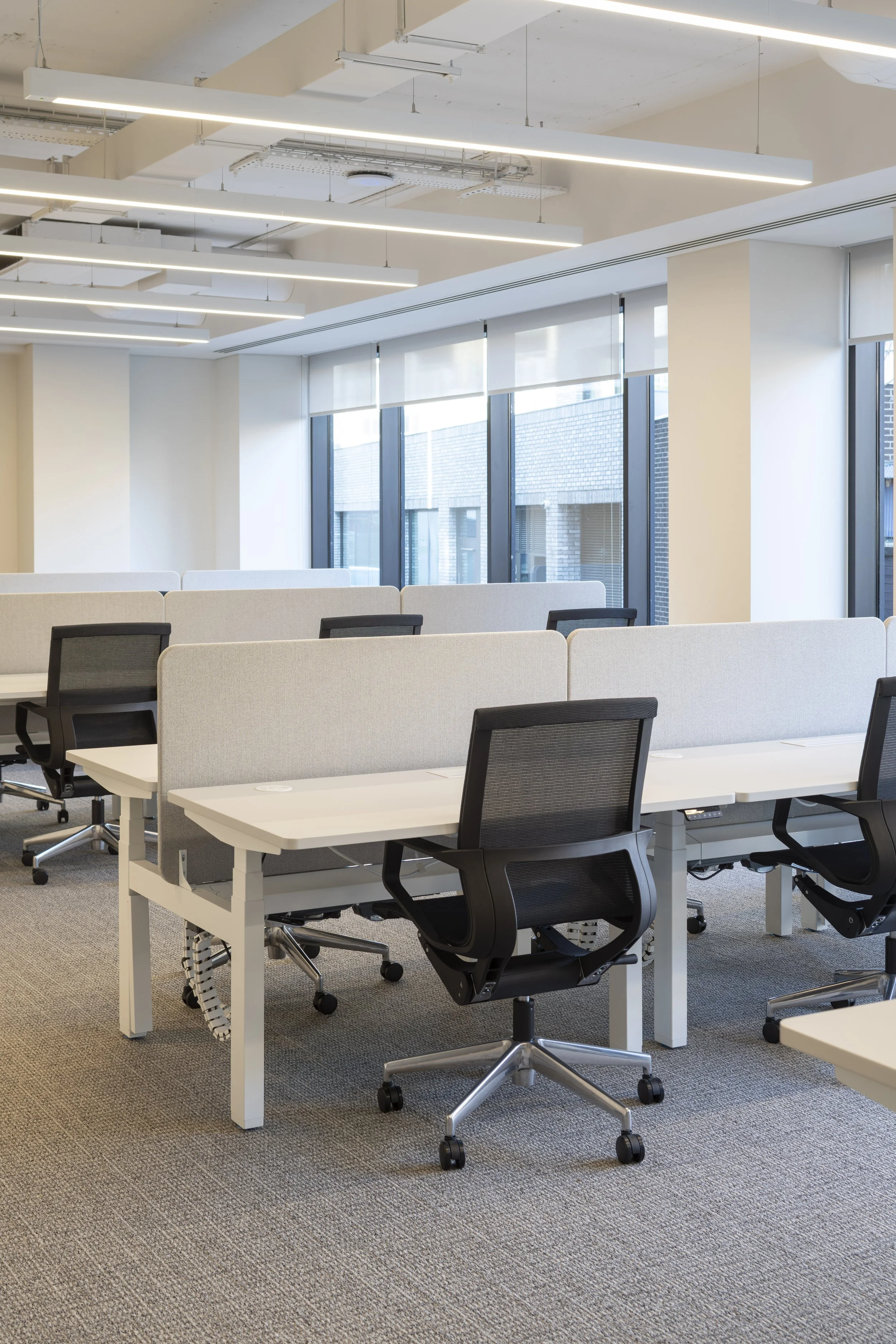 Empty modern office space with white desks, black office chairs, gray partitions, large windows, and ceiling lights.