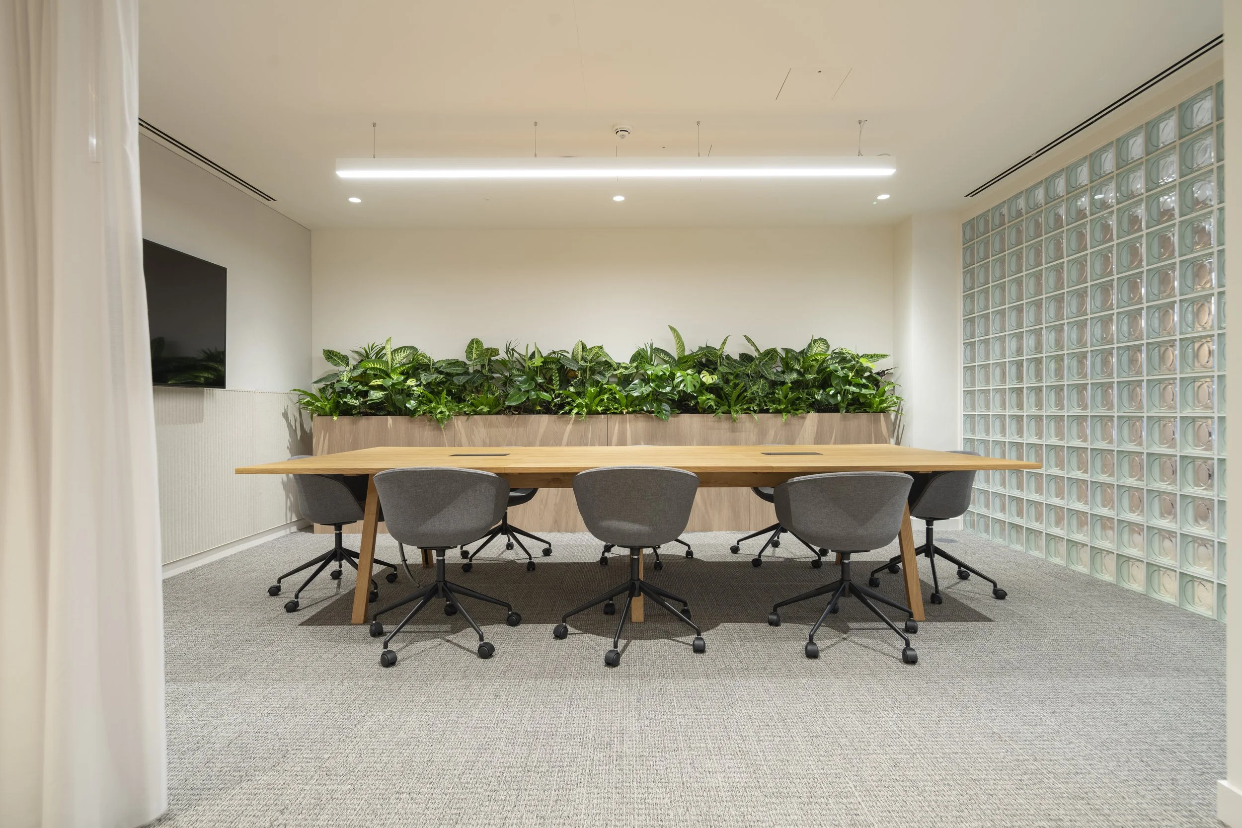 Conference room with a wooden table, gray chairs, a green plant partition, and a glass block wall.