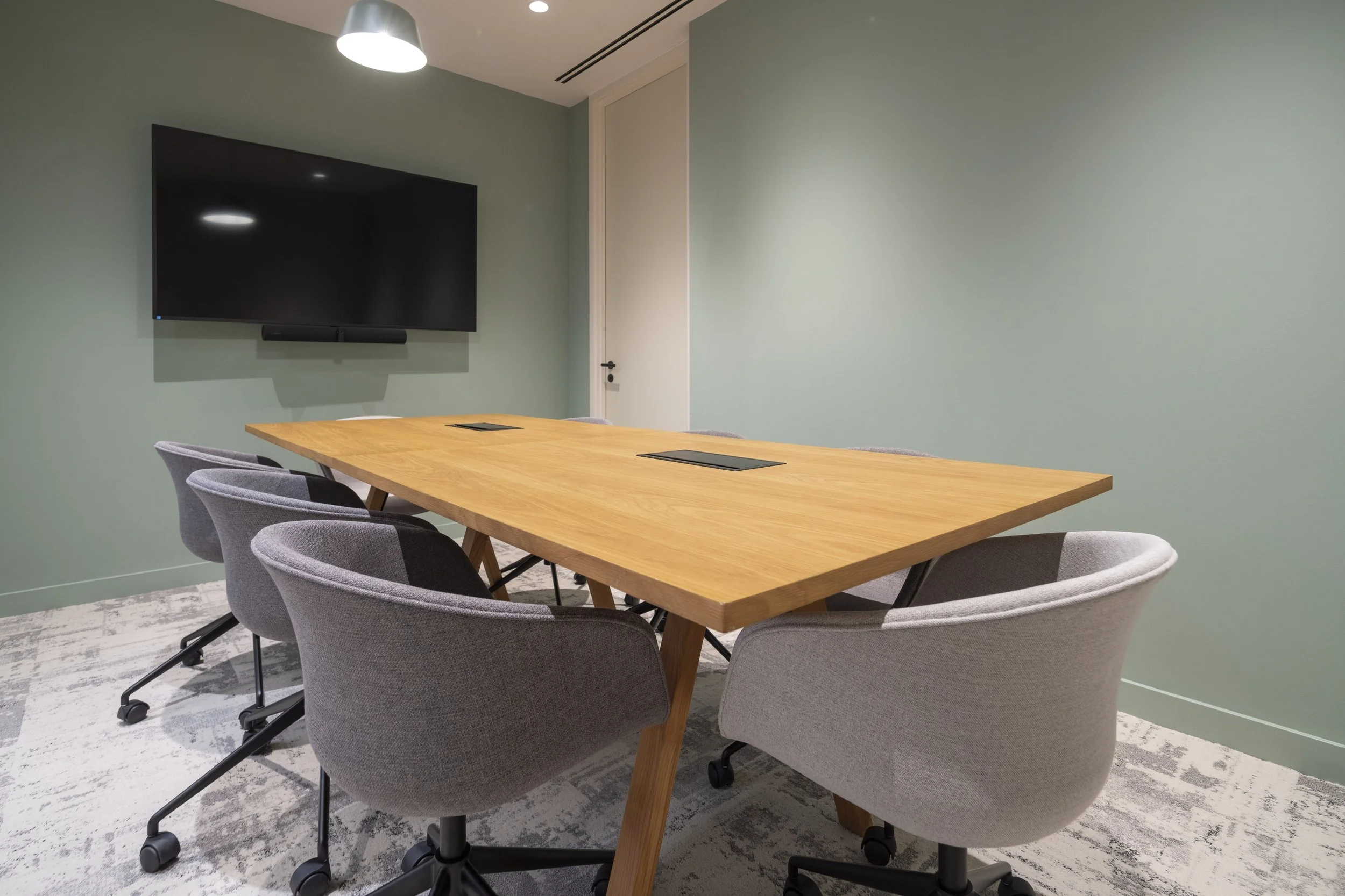 Empty conference room with a large wooden table, six gray chairs on casters, a wall-mounted flat-screen TV, and a closed door.