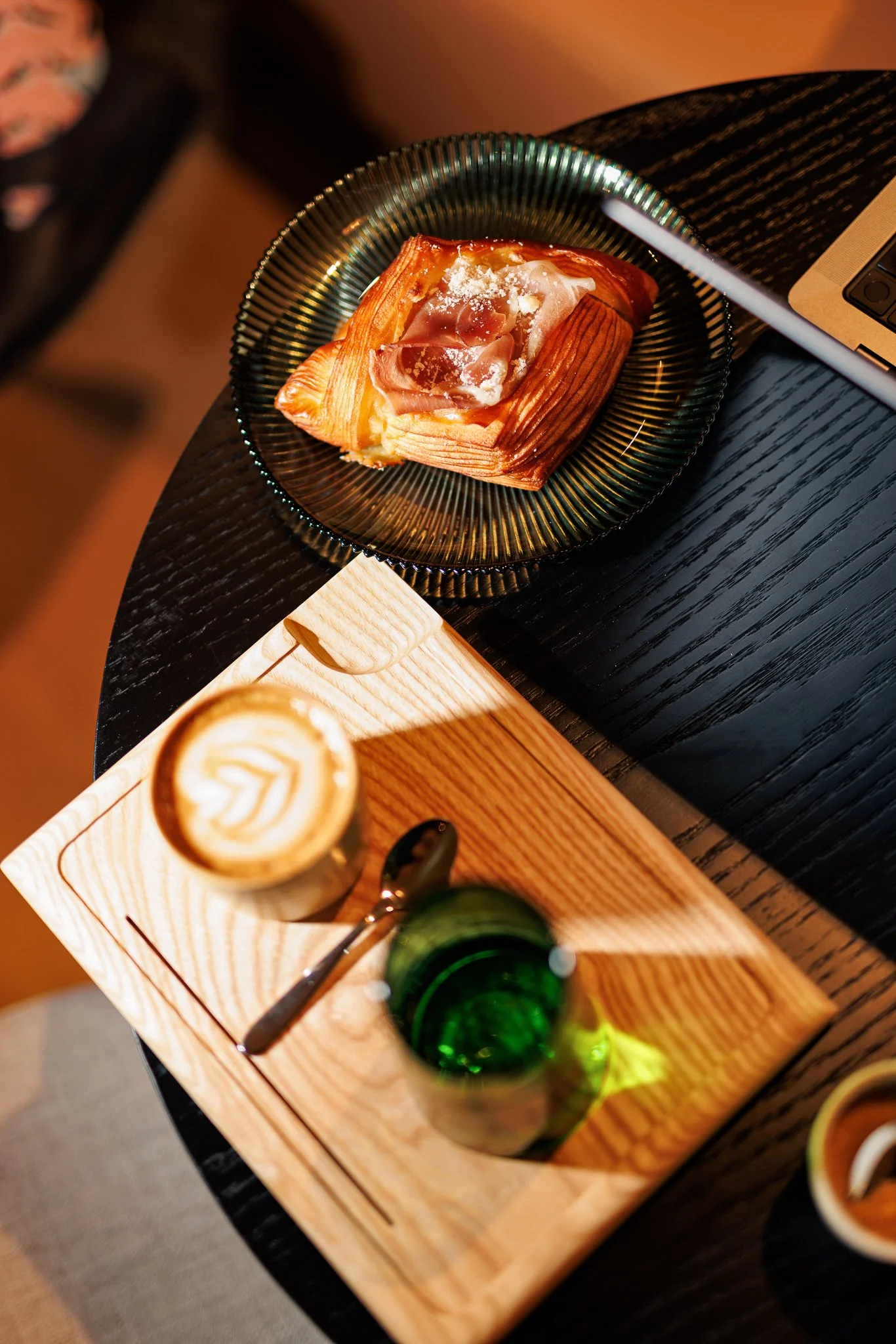 A pastry topped with ham on a black textured plate, a cup of coffee with latte art, and a green glass of water on a wooden tray.