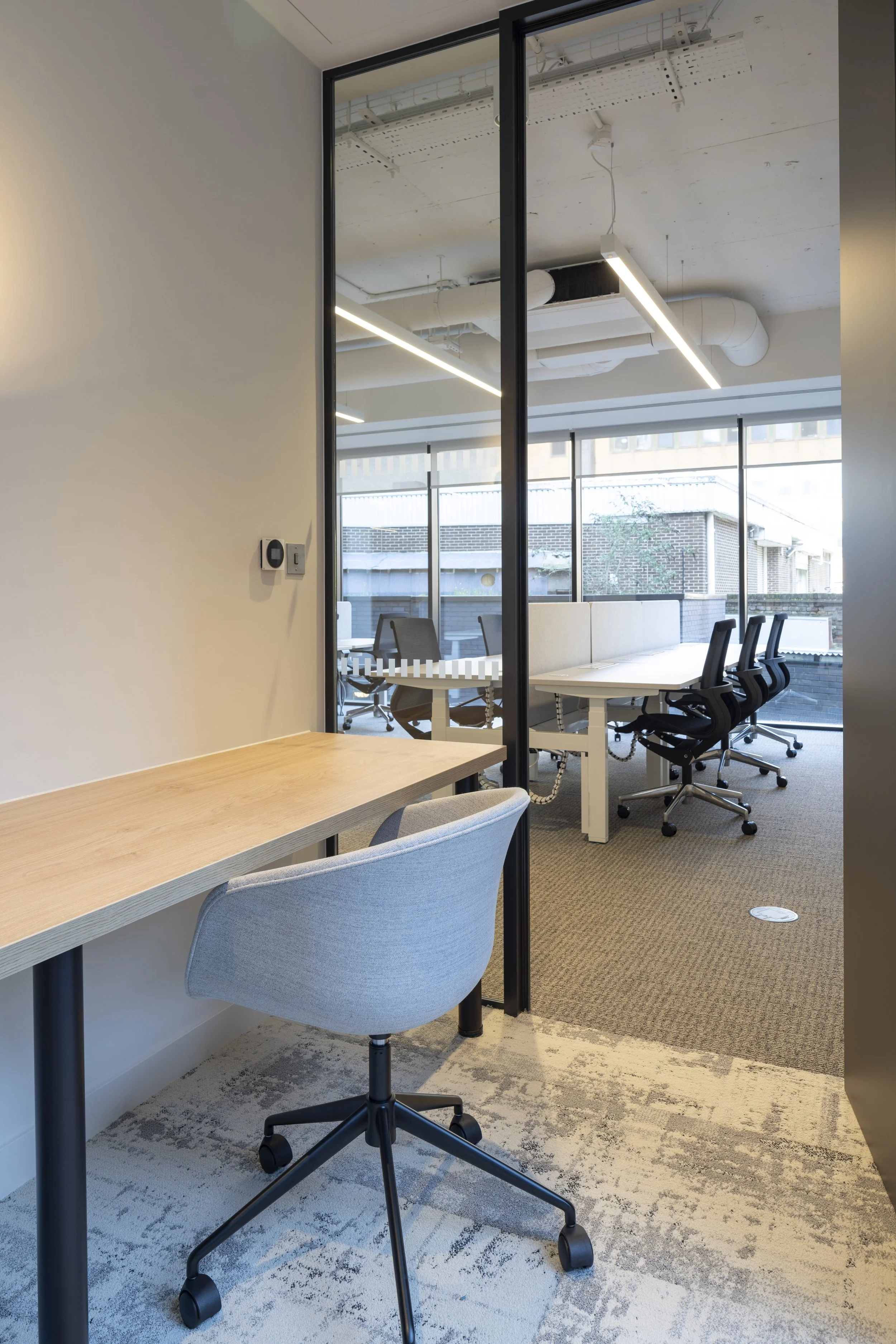 Modern office with a small desk and a light gray rolling chair near a wall, separated by a glass partition from a conference room with white tables and black rolling chairs, large windows, and ceiling lights.