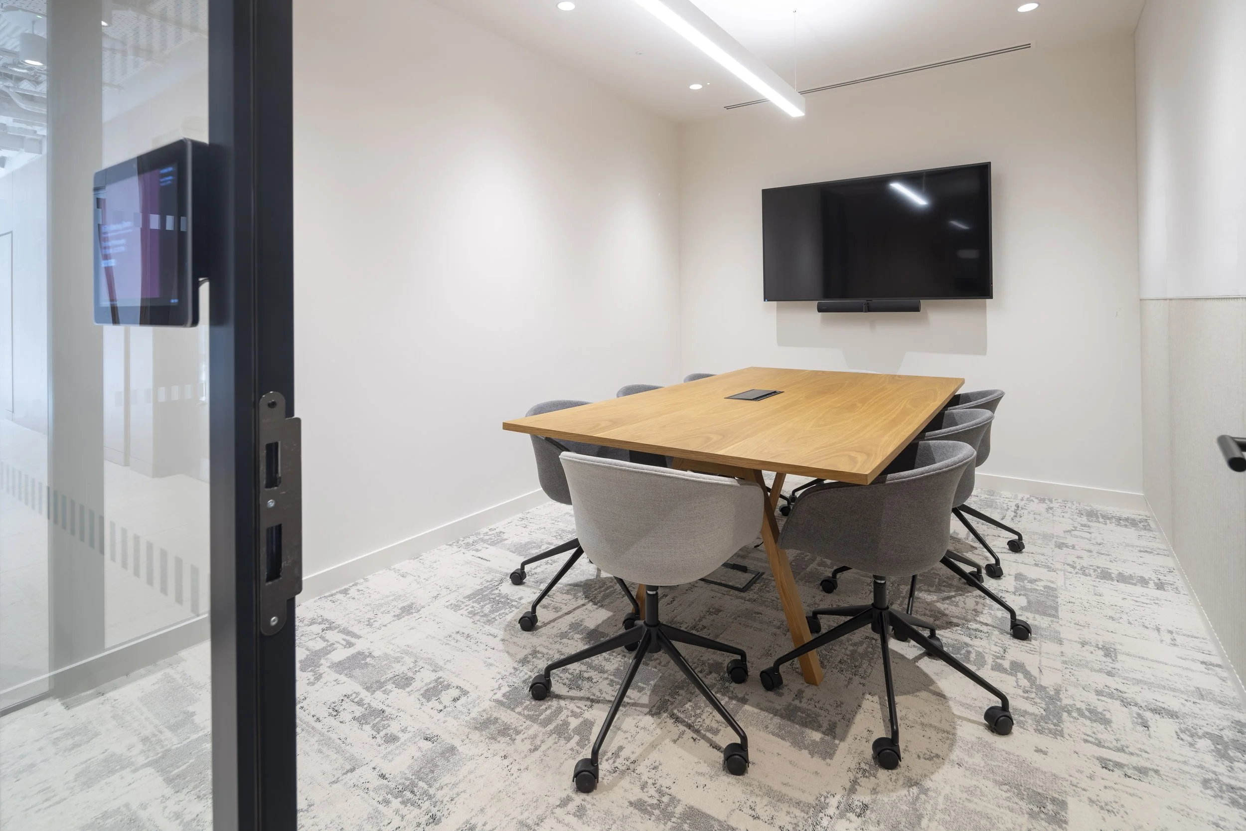 Modern conference room with a wooden table, eight gray swivel chairs, a wall-mounted flat-screen TV, and a wall-mounted speaker. Neutral wall colors and a patterned carpet are also visible.