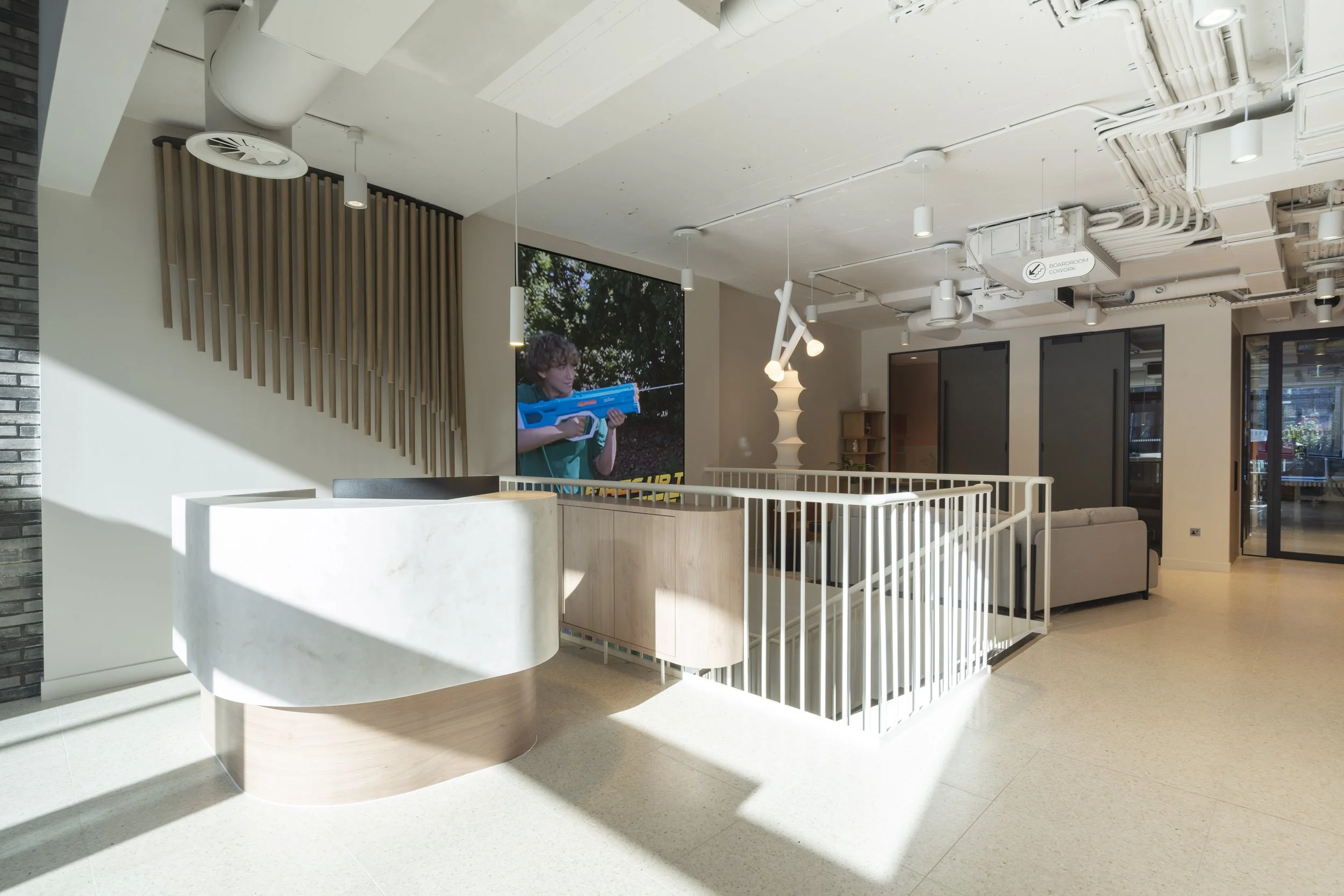 Modern interior space with a reception desk, a large screen showing a person holding a toy gun, and seating area, with sunlight streaming through windows.