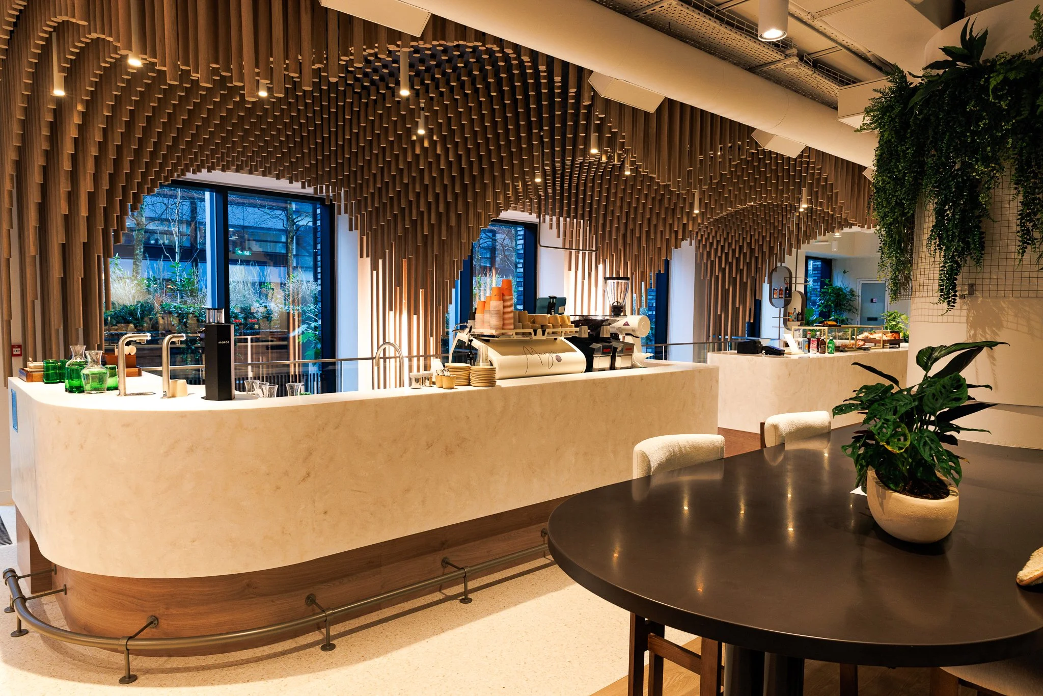 Modern cafe or coffee shop interior with a curved white marble counter, espresso machine, cups, and green glasses. Decorative wooden paneling on the ceiling, large windows, potted plants, and a dark round table with a plant in a white pot.