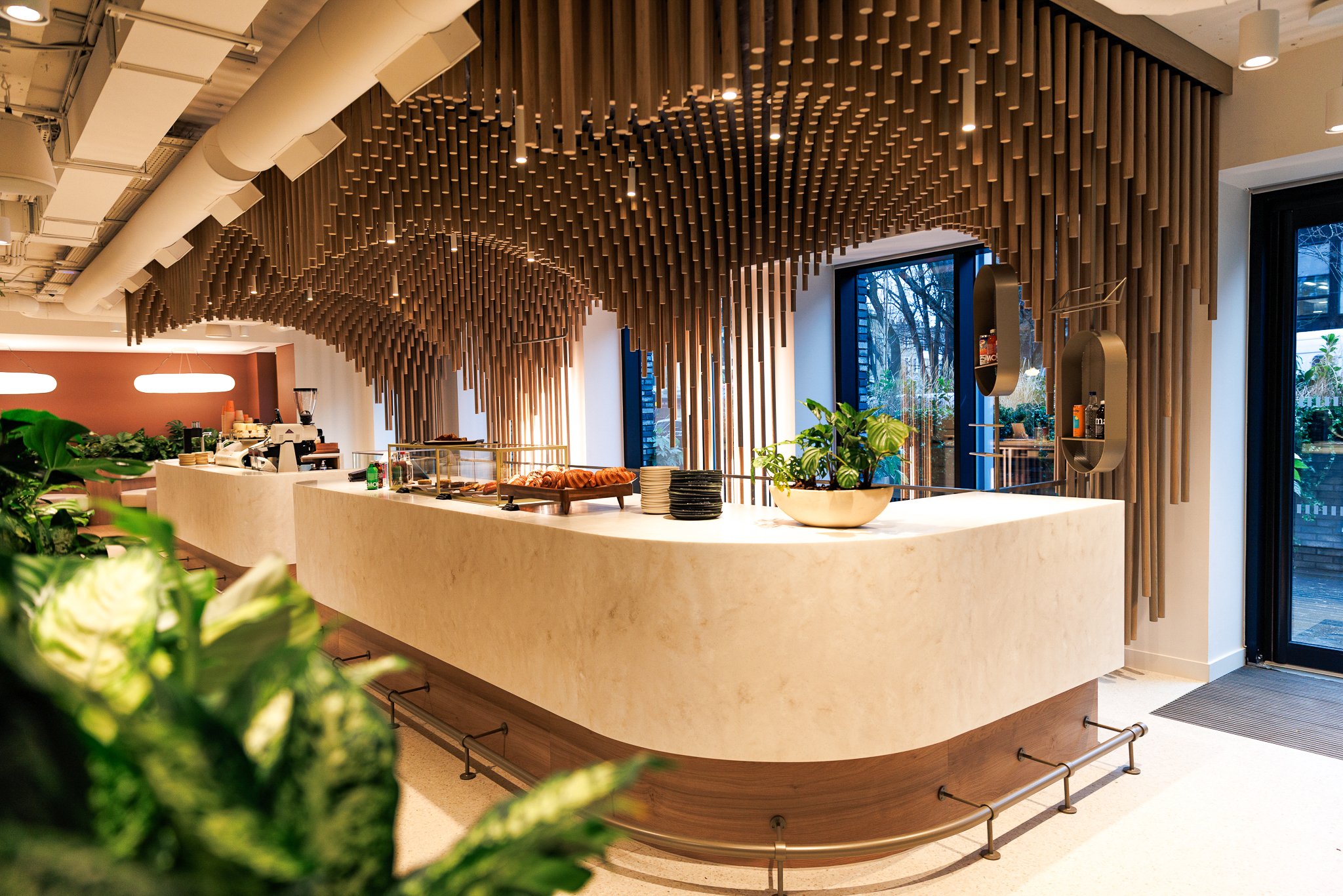 Modern coffee shop counter with a curved white marble surface, potted plants, and a backdrop of wooden vertical slats on the ceiling and wall. Large windows show outdoor trees.