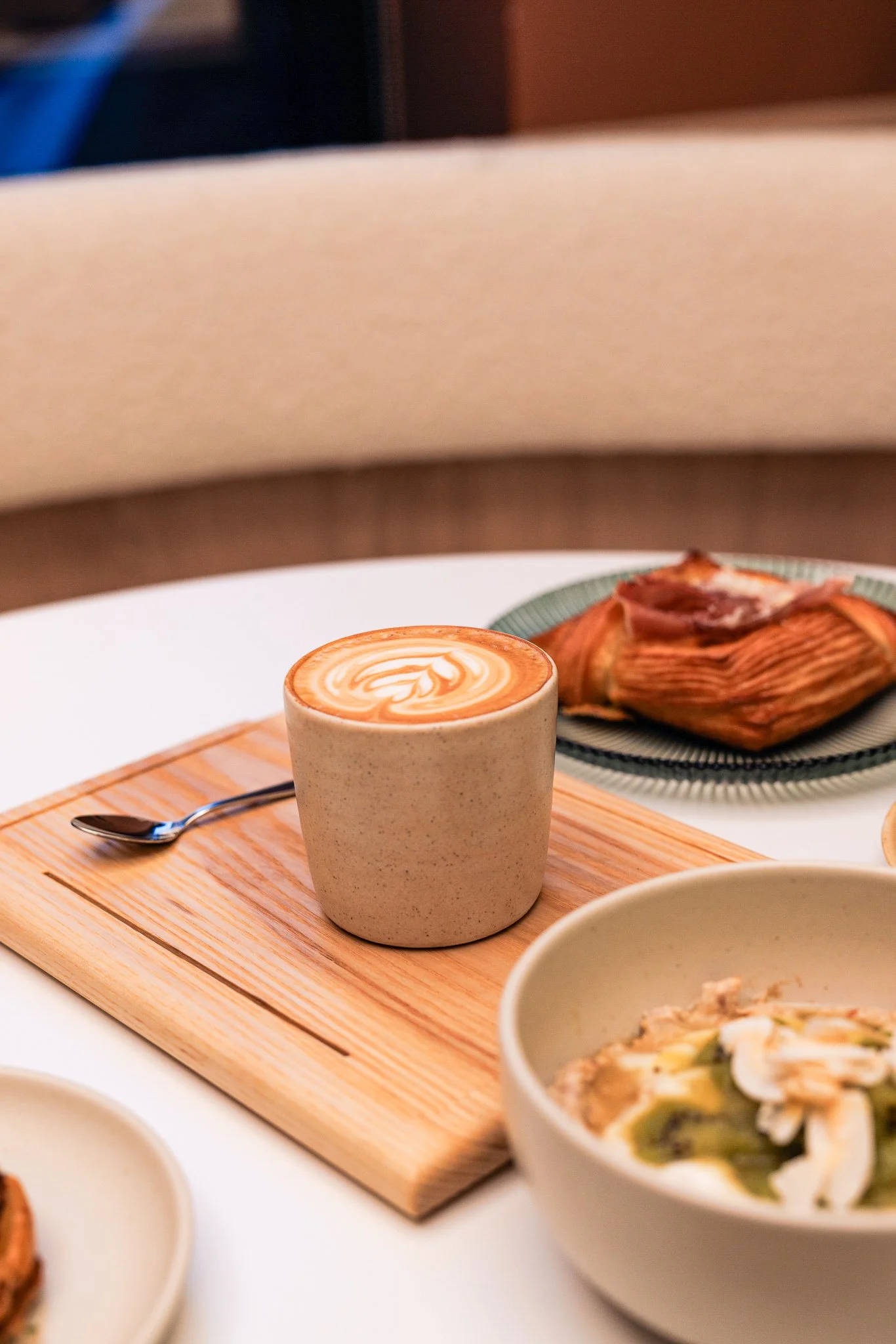 A cup of coffee with latte art on top, placed on a wooden tray on a white table, with a spoon beside it. There are various dishes, including a bowl of salad and a pastry, on the table.