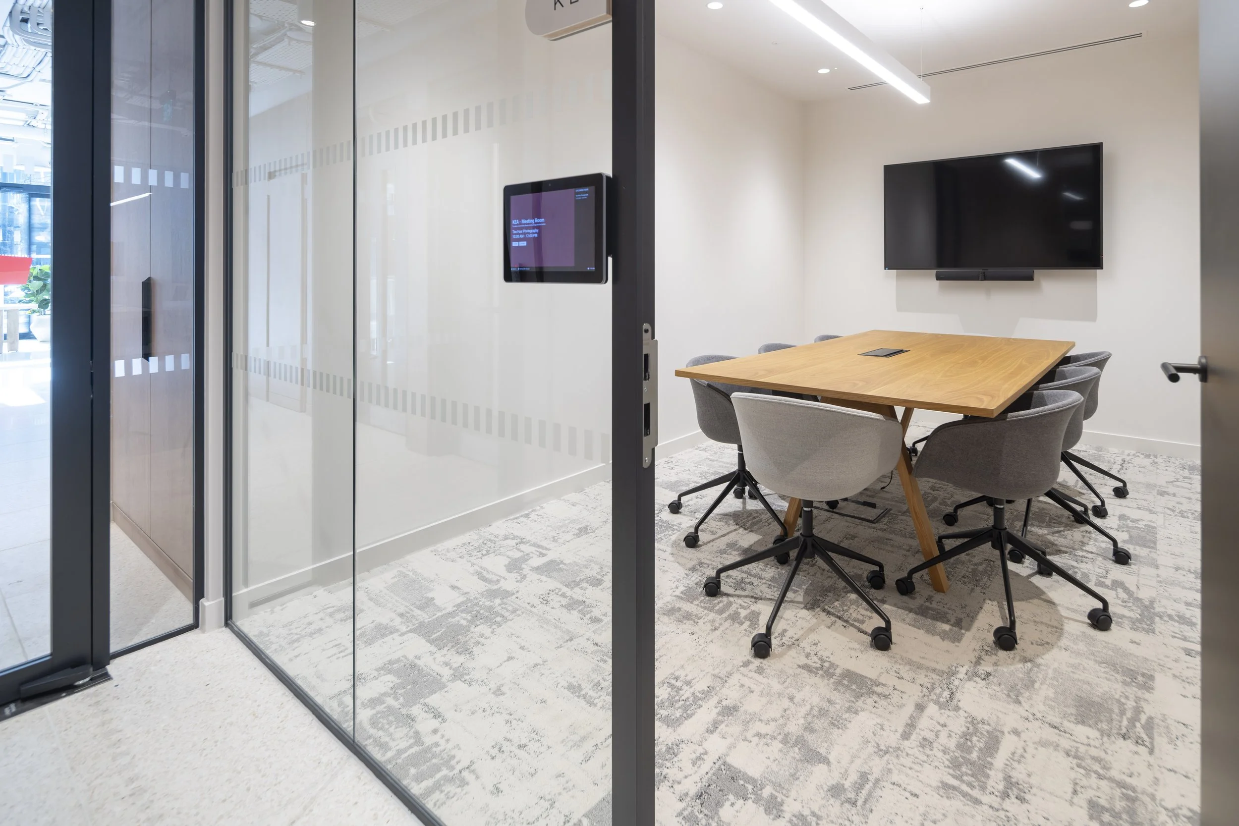 Modern conference room with a wooden table, six gray chairs, a large wall-mounted TV, and a glass wall with digital display.