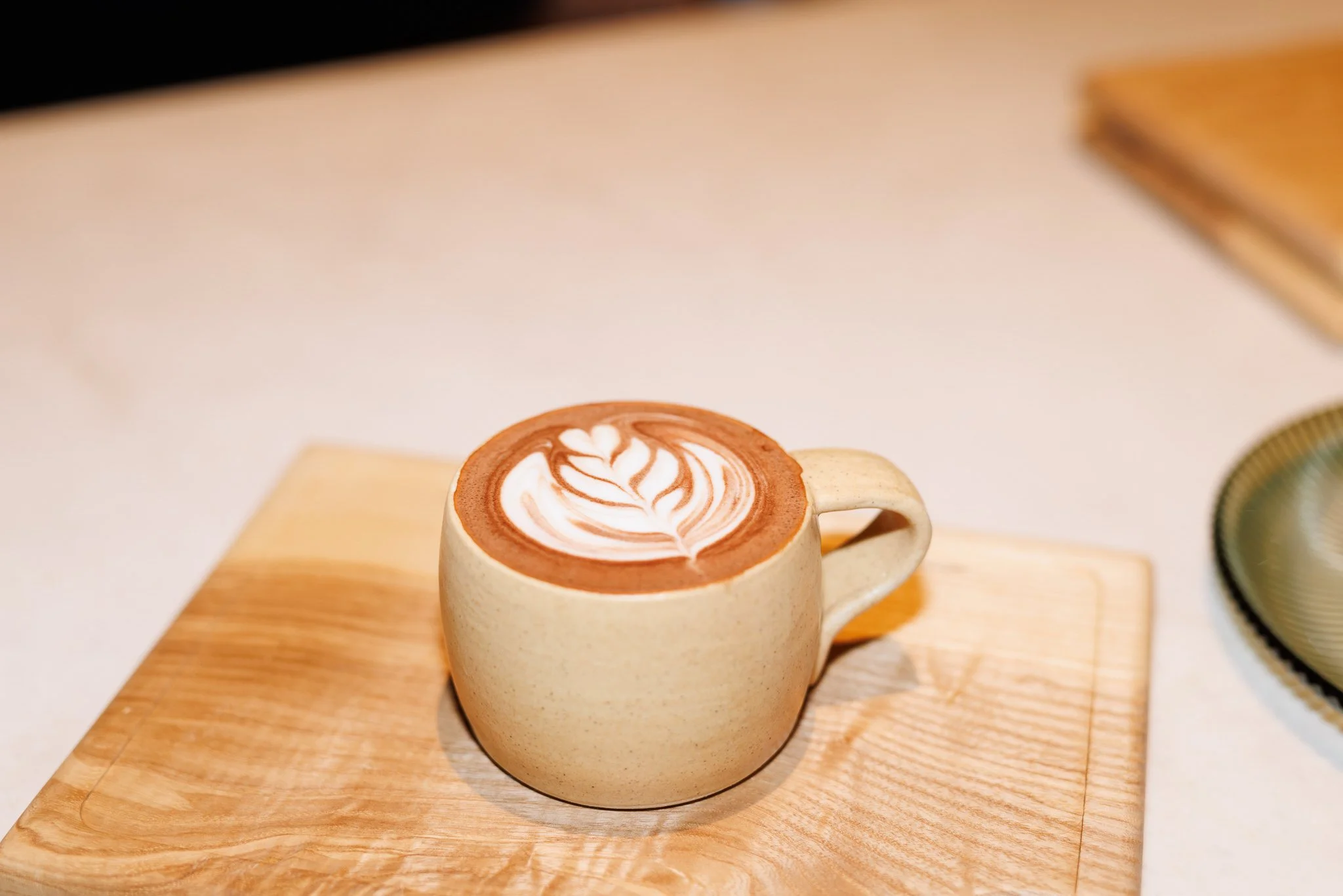 A beige ceramic mug with a latte art design on top, sitting on a wooden tray. The latte art is a rosette pattern with white foam on brown coffee.