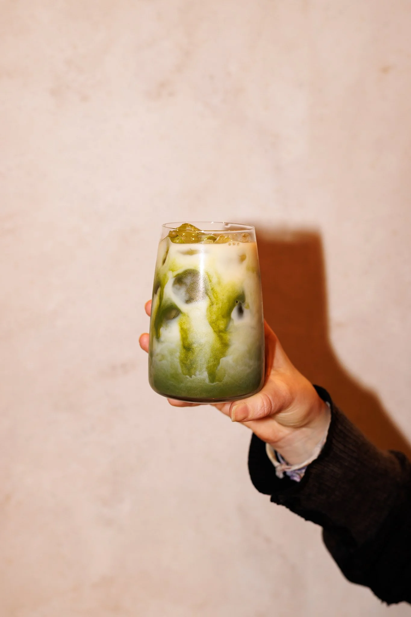 A hand holding a glass of iced matcha latte with green and white swirls against a neutral wall.