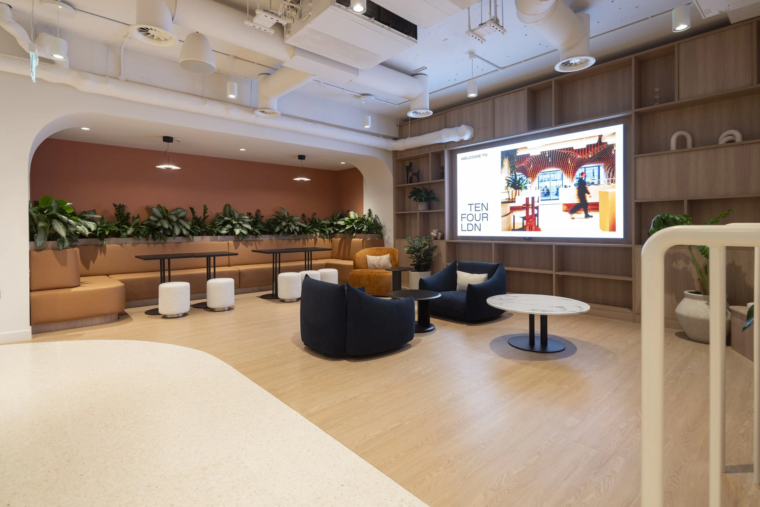 Modern lounge area with black and blue armchairs, small round tables, a marble-top table, a tan booth with plants behind, and a large digital screen on a wooden wall.