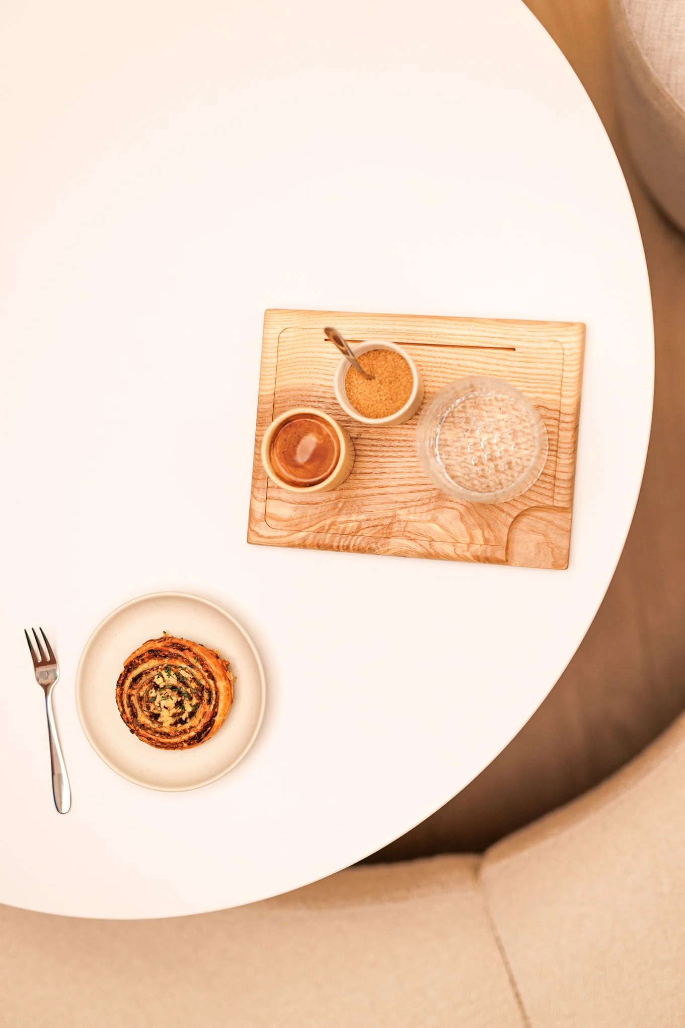 Top-down view of a white round table with a plate holding a cinnamon roll and a fork, along with a wooden tray containing a glass of water, a small bowl of sugar, and a small bowl of brown sugar with a spoon.