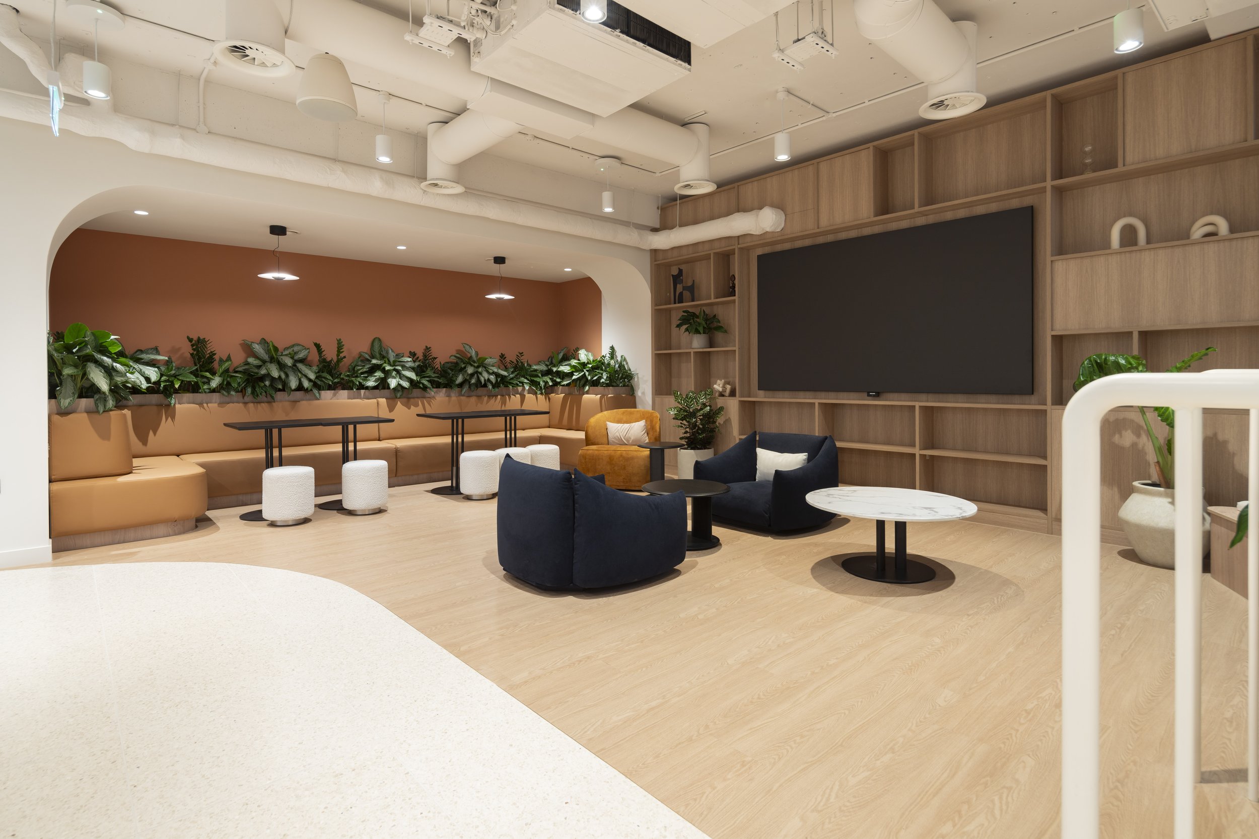 Modern lounge area with navy blue chairs, round marble tables, potted plants, and a large television on a wooden shelving wall.