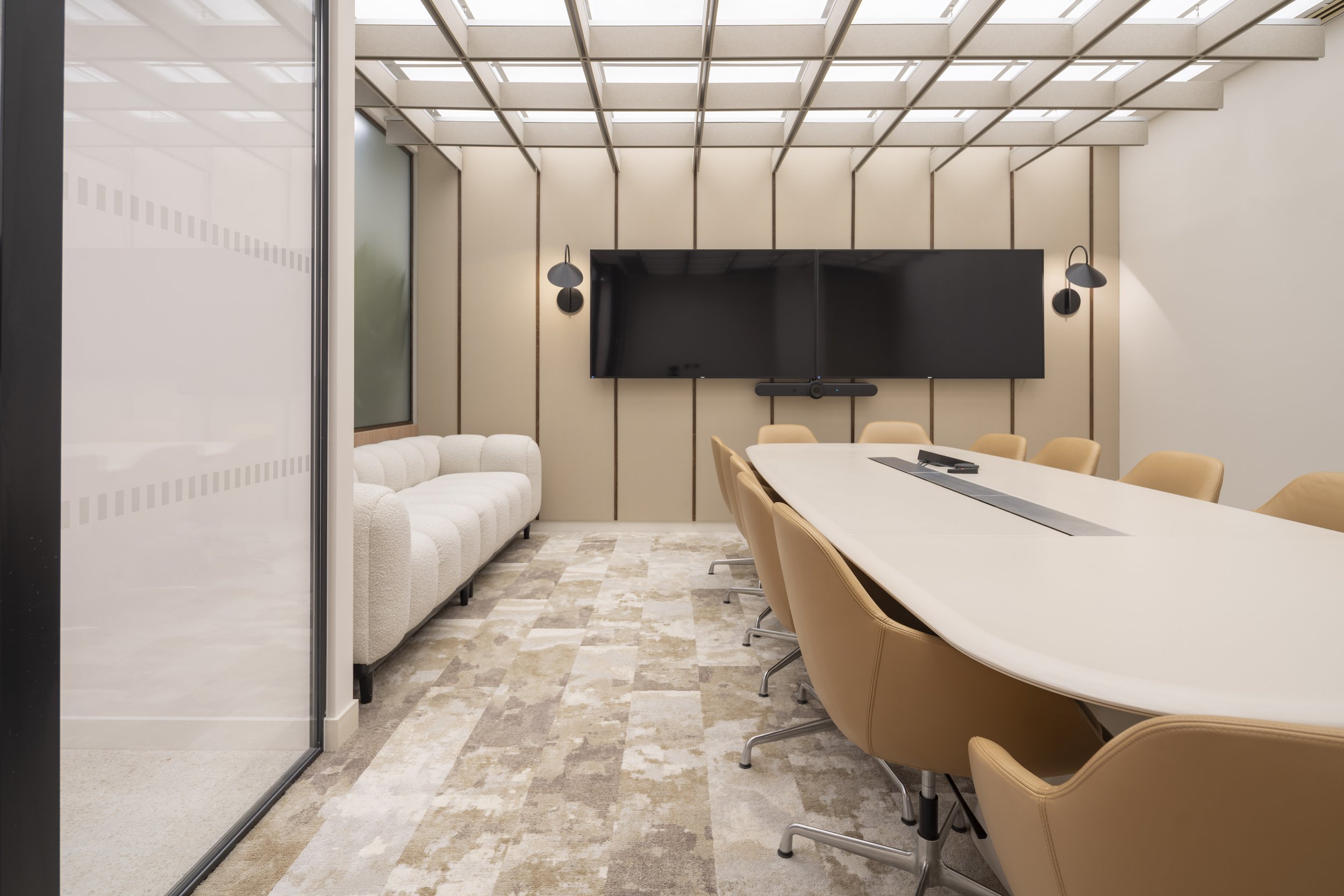 A modern conference room with a long white table surrounded by tan chairs, a large black screen on the back wall, a white sofa on the left, wall-mounted lights, a glass wall on the left side, and a skylight ceiling letting in natural light.