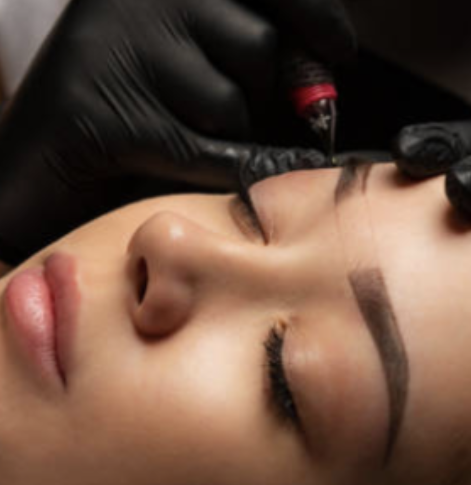 A person lying down with eyes closed, getting a cosmetic tattoo or permanent makeup procedure on eyebrows while a technician with black gloves applies ink with a small tool.