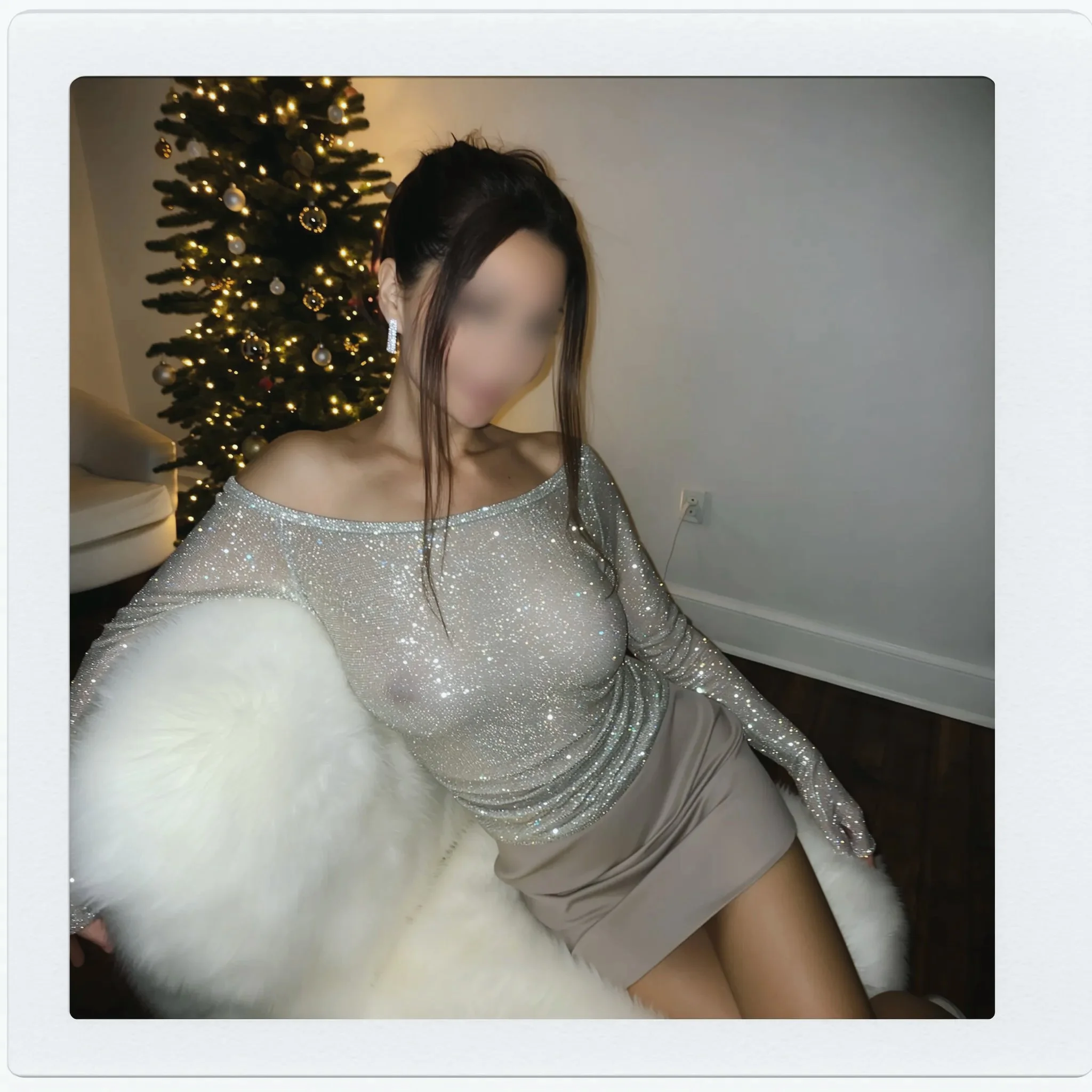 A woman in a glittery silver off-shoulder top and beige skirt sitting on a white furry chair. Background features a decorated Christmas tree with lights and ornaments.