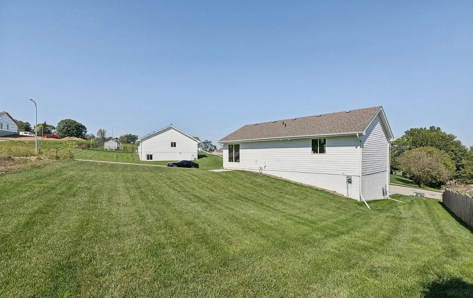A suburban backyard with a large, well-kept grassy lawn, a white house with a brown shingled roof, a driveway with a black car, and other houses in the background under a clear blue sky.