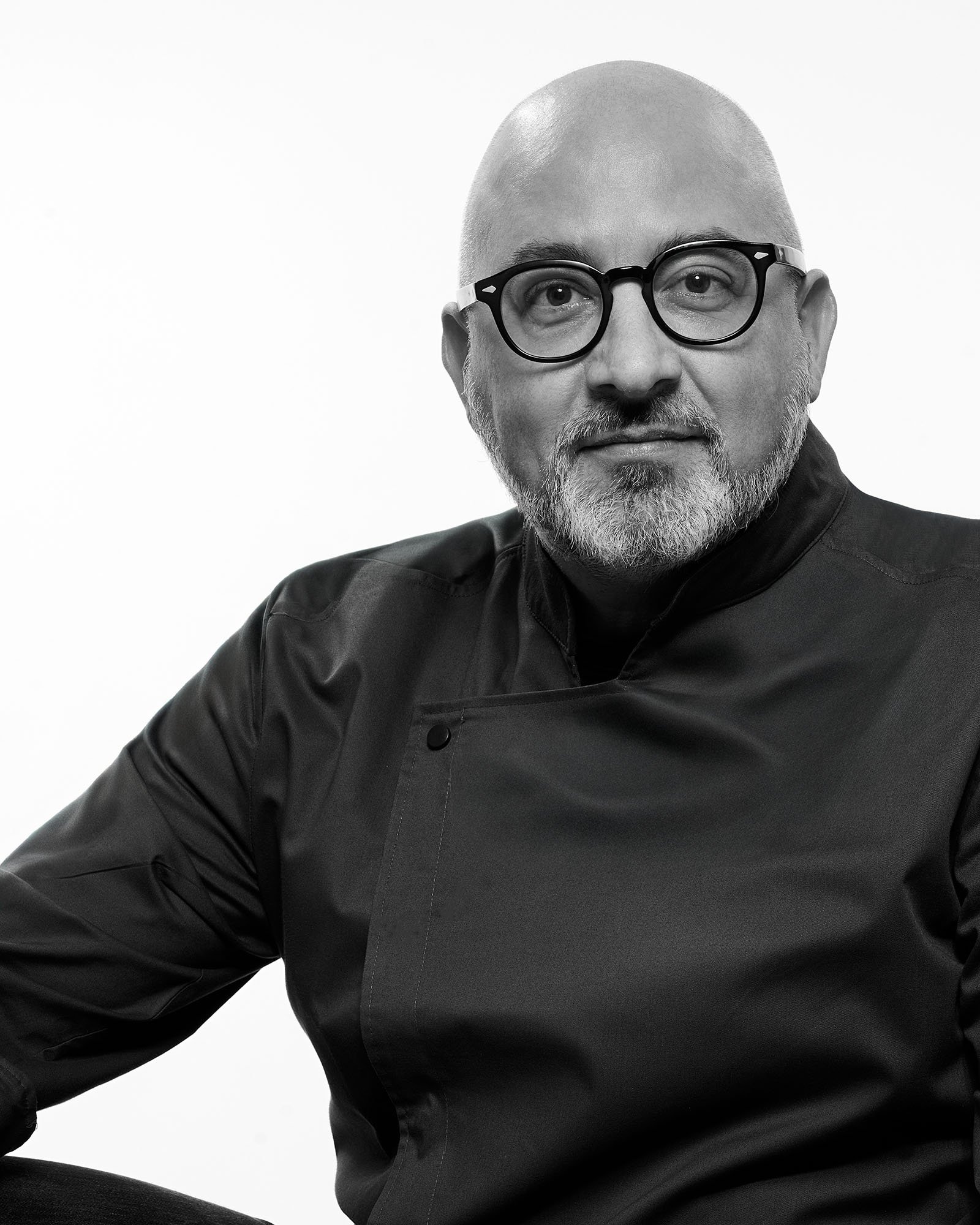 Uomo con barba e occhiali rotondi, vestito da chef nero, che sorride leggermente