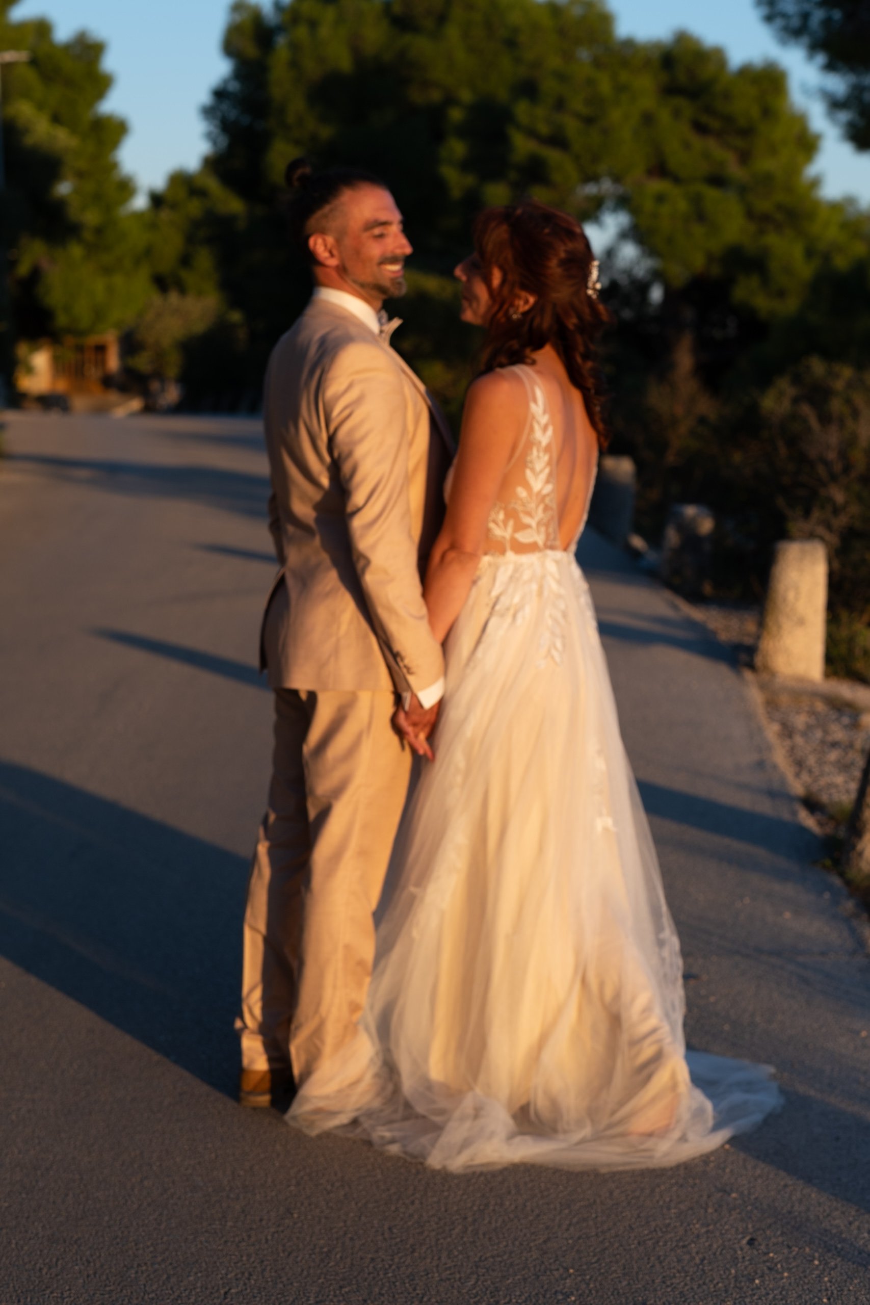 Bride and groom standing on road, holding hands, wearing wedding attire, outdoor setting with trees.
