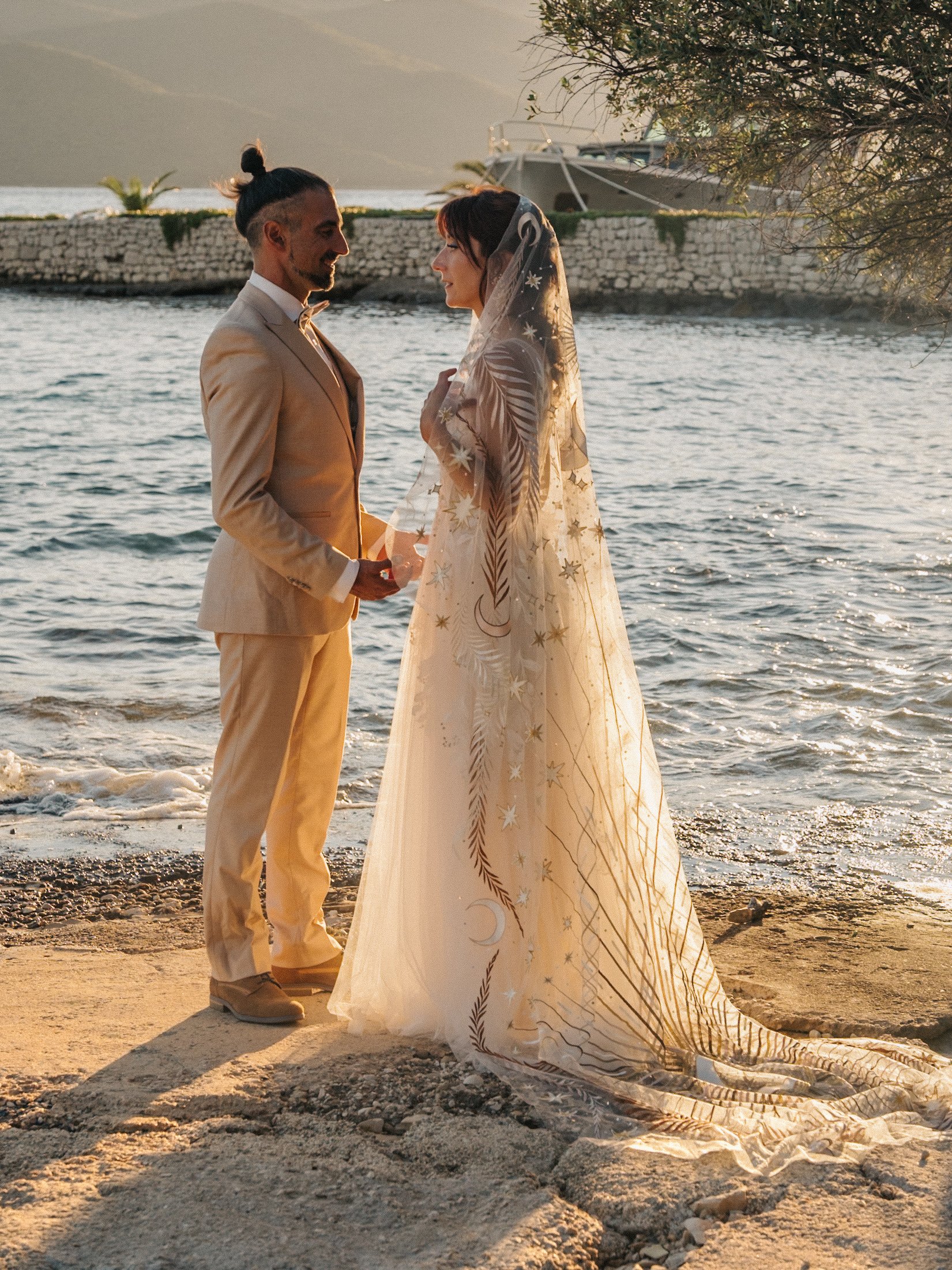 Bride and groom standing by a lakeside, sunset lighting, wearing elegant wedding attire, with the bride in a long veil.