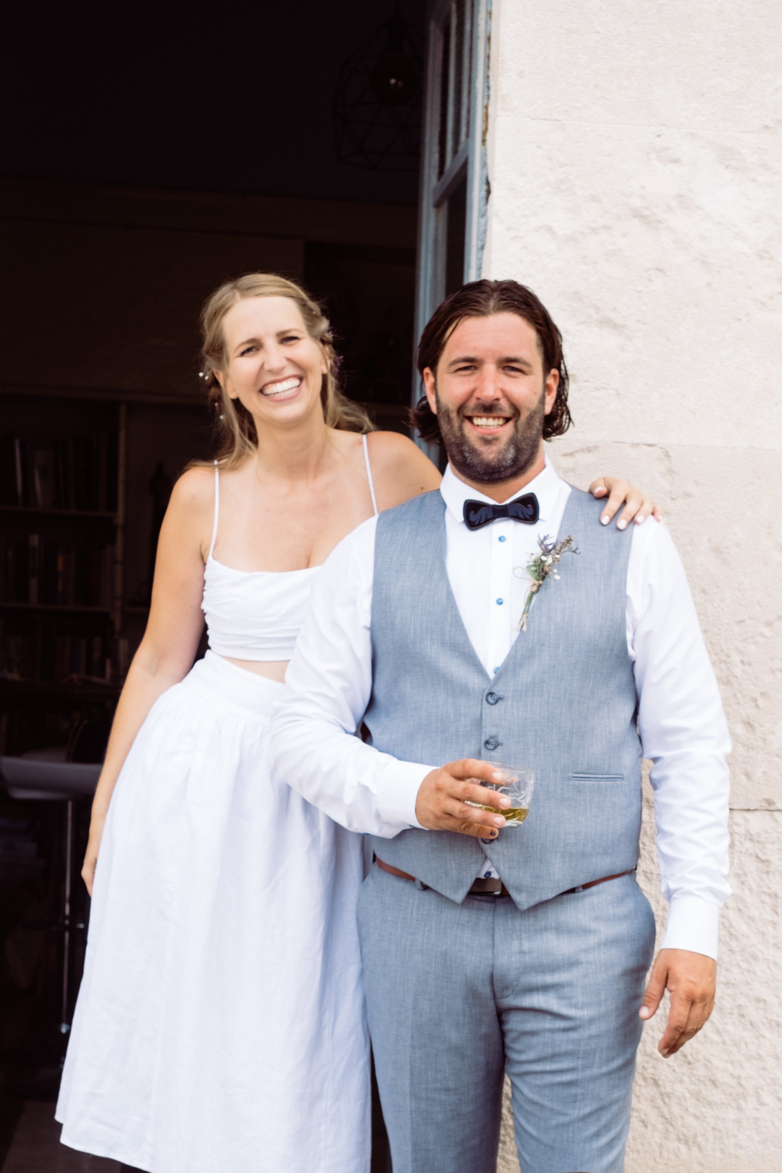 Smiling couple, woman in white dress, man in gray suit and bow tie, standing outside a building.