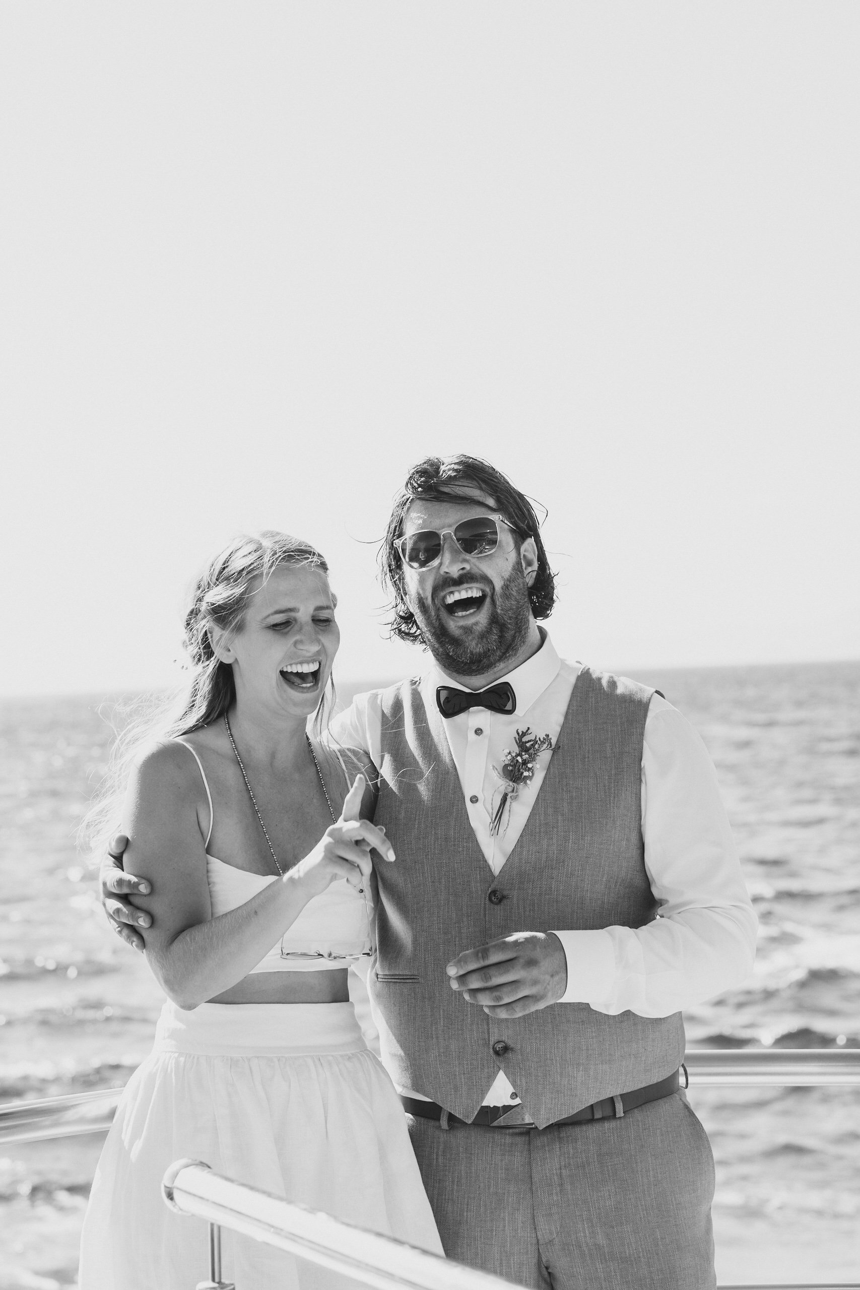 Happy couple laughing on a boat, dressed in wedding attire with ocean in the background.